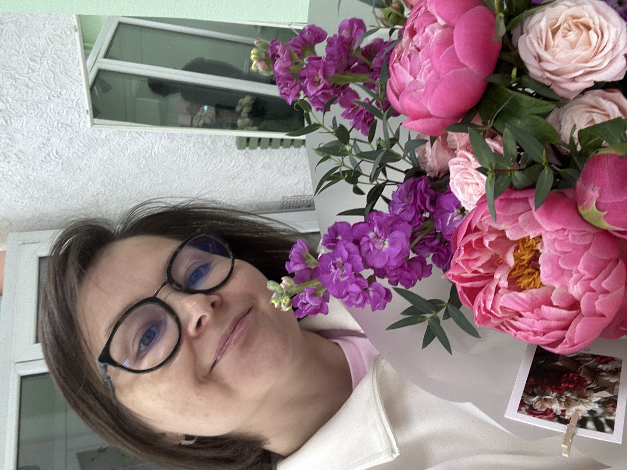 A woman with brown hair and glasses is holding a bouquet of pink and purple flowers. The bouquet also has a small photo attached with a clothespin. The background includes a window and a wall with patterned wallpaper. This image might be taken in Izhevsk, Russia, but there is no specific visual information to confirm the location.