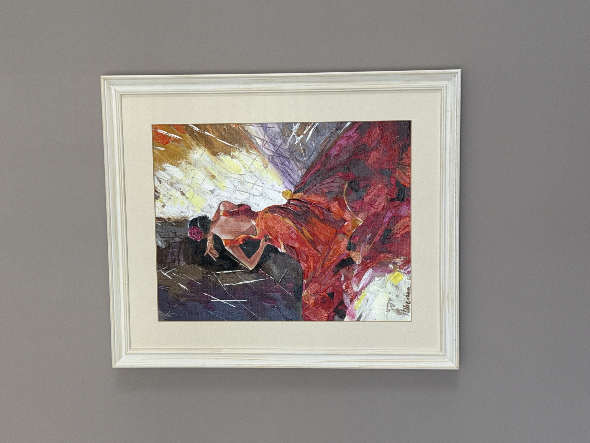 A framed oil painting of a flamenco dancer in a red dress is displayed on a grey wall. The painting, done in an impressionistic style, features bold brushstrokes and vibrant colors, primarily reds and yellows, with dark accents. The dancer, with her back to the viewer, is depicted in mid-motion, her voluminous red skirt swirling around her. She has dark hair adorned with a red flower and her left arm is extended. The background is a chaotic blend of dark and light colors, suggesting dramatic lighting. A cream-colored mat surrounds the painting, and it is encased in a distressed white wooden frame. The painting is signed in the lower left corner with what appears to be "Villadema". This artwork is located in Burgas, Bulgaria, specifically in the City Art Gallery "Petko Zadgorski", which houses a significant collection of Bulgarian art, including impressionist works.
