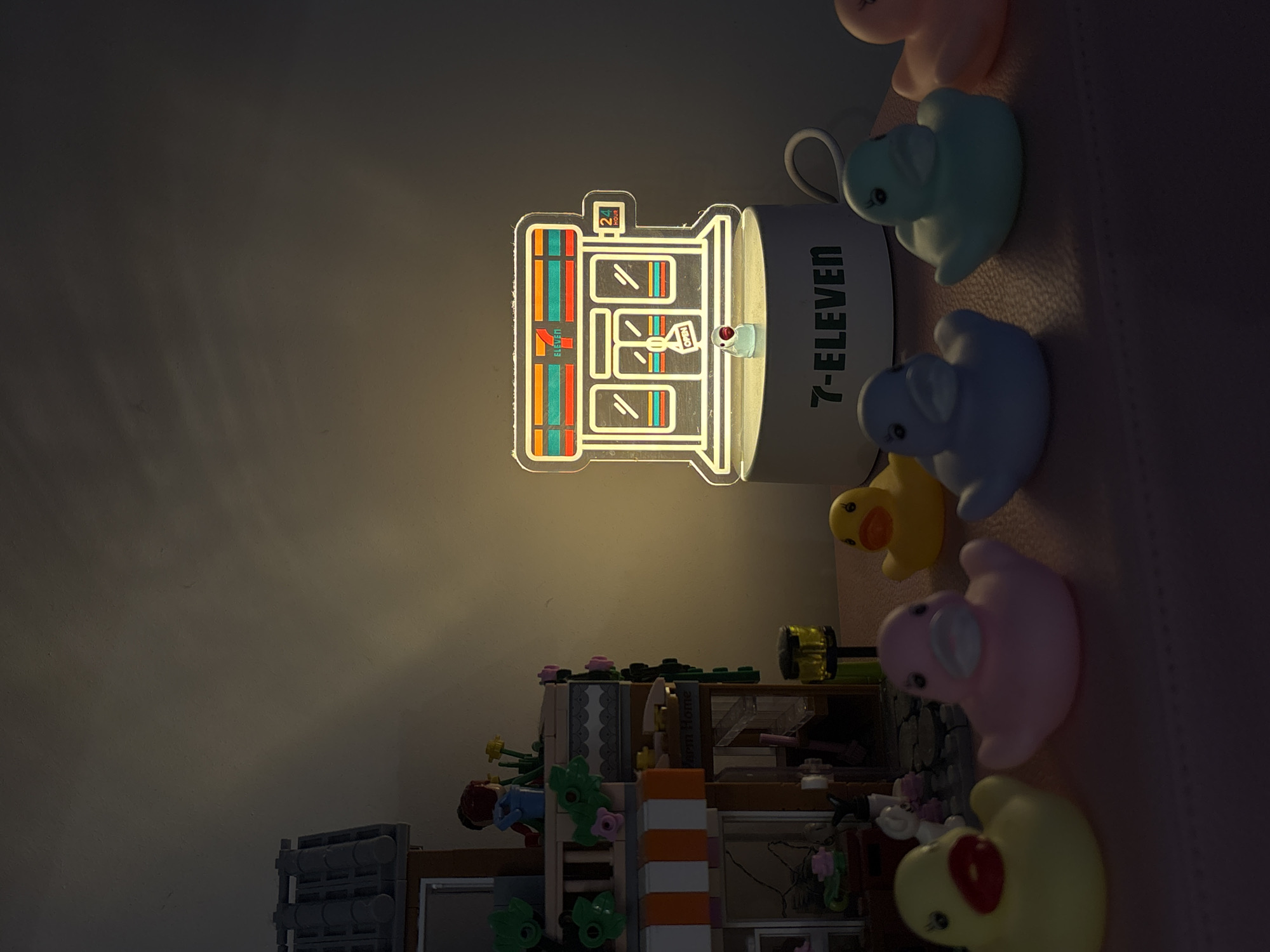 This dimly lit, static image captures an indoor scene, likely at night, featuring a decorative arrangement on a surface against a plain wall. The central focus is a glowing desk lamp designed as a miniature representation of a 7-ELEVEN convenience store. The lamp is made of translucent material, outlining the building with bright lines and showing colorful shelves inside. Text visible on the lamp's design includes "7-ELEVEN", "24 HOUR", and "OPEN". The cylindrical base of the lamp also prominently displays "7-ELEVEN" vertically.
To the right of the lamp, a row of several rubber ducks of various pastel colors is arranged, including pink, light blue, dark blue, and yellow, with one light yellow/greenish duck also visible. Below the lamp, a detailed miniature structure, resembling a house or building made from interlocking plastic blocks (like LEGO), is present. This structure features walls, windows, and small plant decorations in green and pink. Faint text reading "Farm Home" can be seen on one of the block components. The scene suggests a personal collection or display. The provided location context is Krasnoyarsk, Russia.