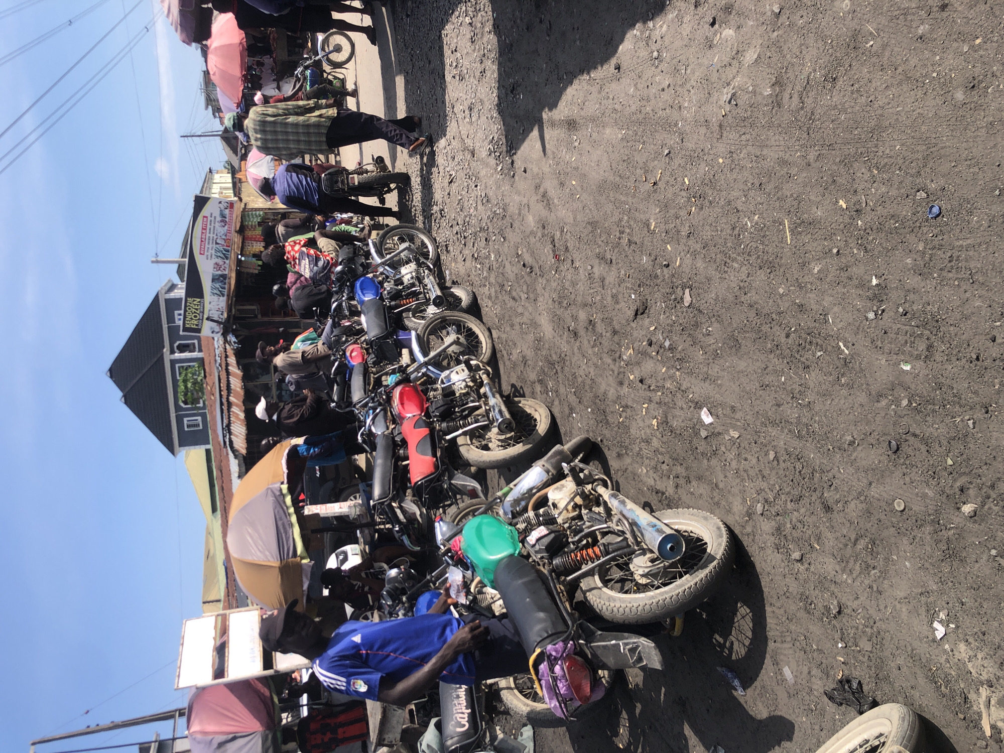 In Ado, Nigeria, a dusty, unpaved market street is crowded with motorcycles, likely serving as taxis. The scene is bright and sunny, with a clear blue sky overhead. A man in a blue shirt and black cap stands beside a green-tanked motorcycle, seemingly waiting for a customer. Behind him, a row of motorcycles stretches down the street, some with red seats, others with black. In the background, market stalls and buildings line the street, some with colorful umbrellas offering shade. People can be seen milling about, some seated on motorcycles, others standing. The overall impression is of a busy, working market day.