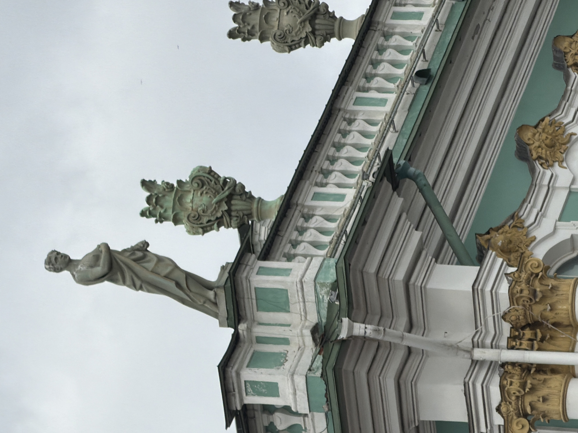 The image depicts architectural details of a building in Saint Petersburg, Russia. A statue of a human figure is visible, serving as a decorative element. Other prominent elements include ornate carvings, a decorative frieze, and architectural moldings. A drainpipe runs along the side of the building. The sky is overcast.