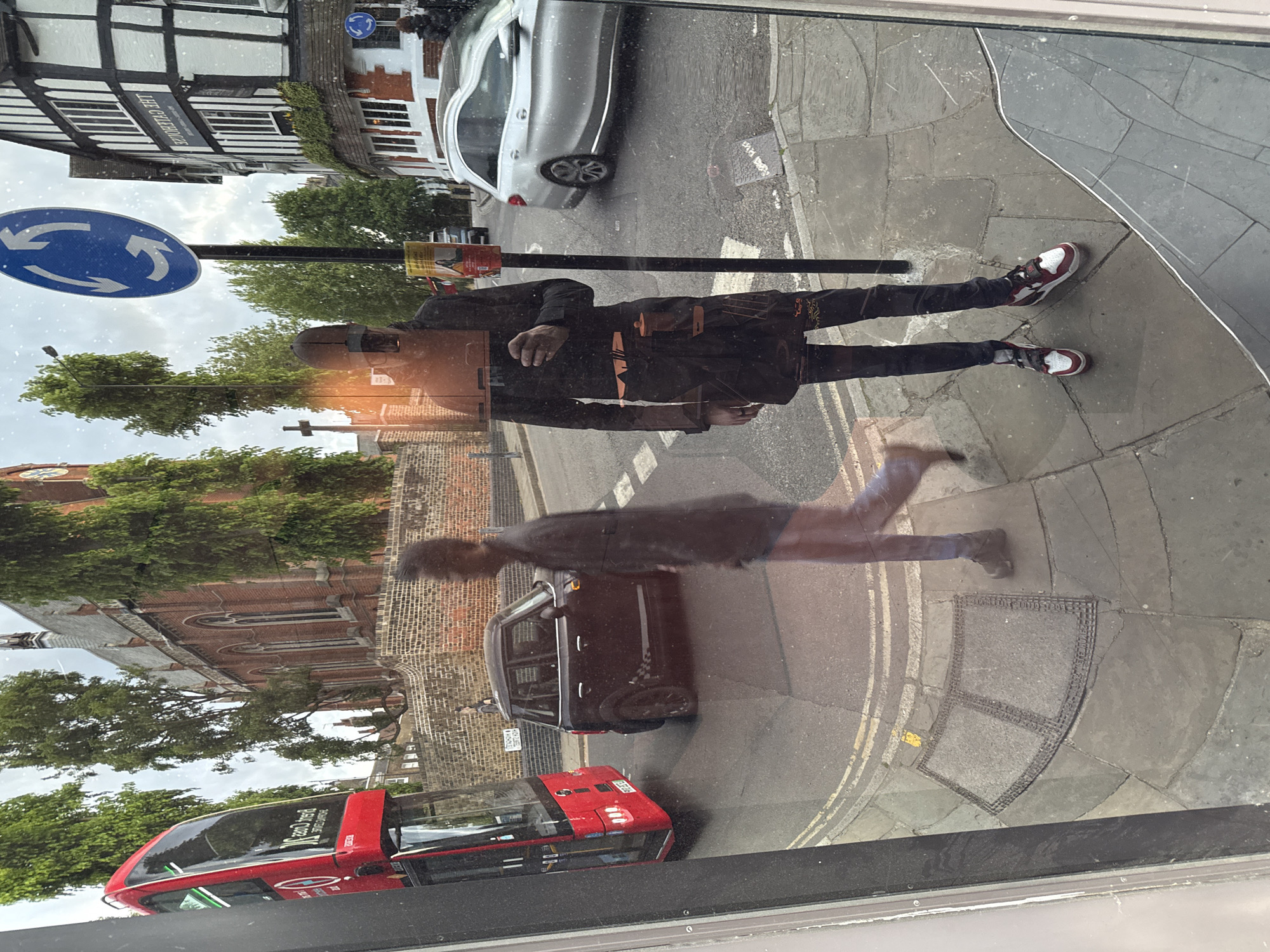 The image is a reflection in a window of a street scene in London, United Kingdom. In the reflection, a person wearing a black coat, black pants, and red and white sneakers is standing on the sidewalk. A pedestrian is walking blurred by the motion and is walking towards the left side of the image. A red double-decker bus, a black car, and a silver car are visible on the street. A blue roundabout sign is mounted on a pole. Buildings, including a half-timbered building, and trees line the street. A church can be seen in the background. The reflection suggests the viewpoint is from inside a building looking out onto the street.