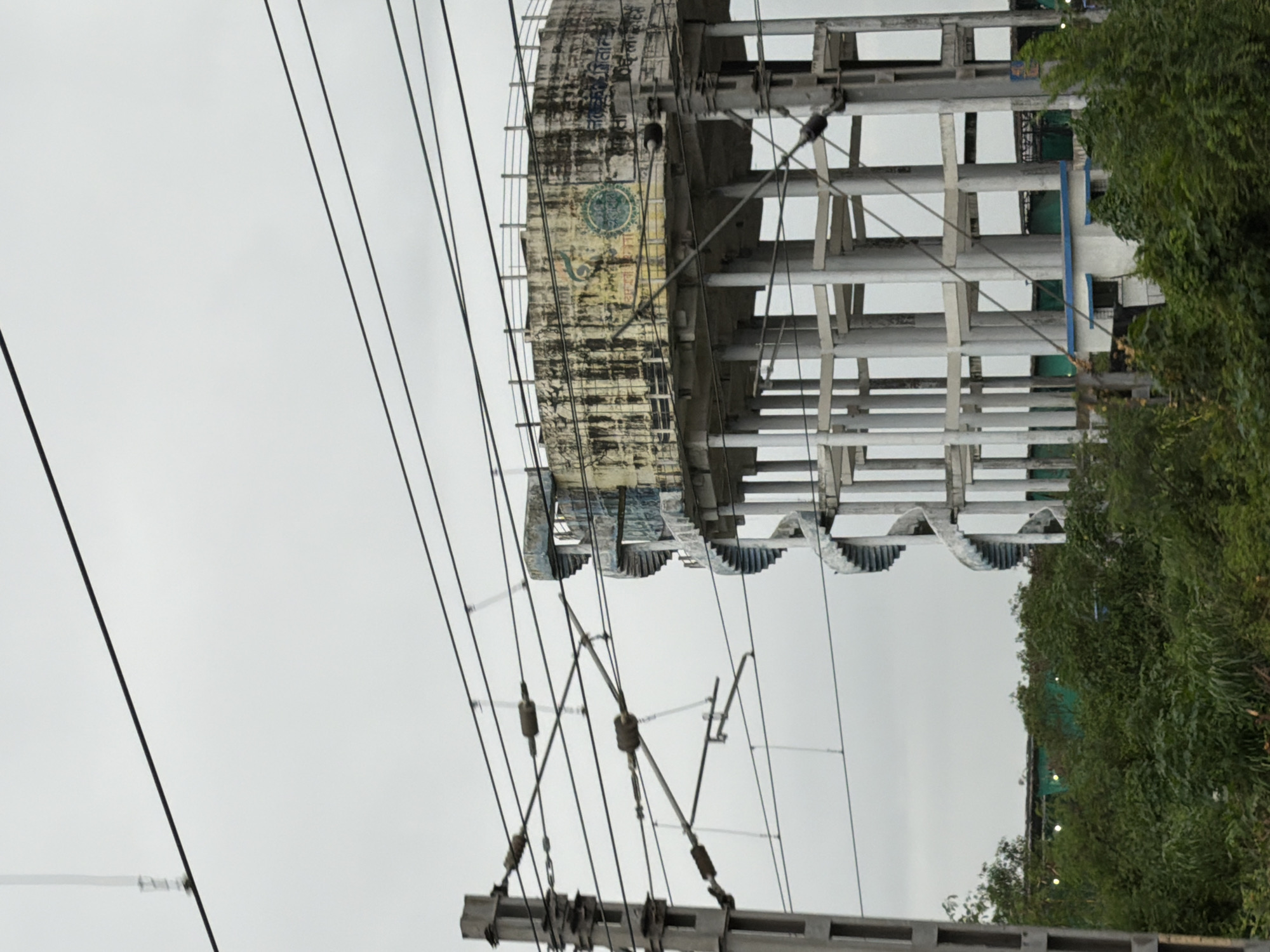 The image shows a tall concrete water tank with a spiral staircase on its side, located in Mira-Bhayander, India. The water tank is supported by several concrete pillars and has some writing and symbols on its surface, which appear to be in Hindi. Overhead power lines for trains crisscross the frame, indicating the tank's proximity to a railway line. The foreground features lush green trees and foliage, suggesting a natural or semi-rural setting within the city. The sky is overcast, contributing to a subdued and slightly muted color palette. There are no people visible in the image.