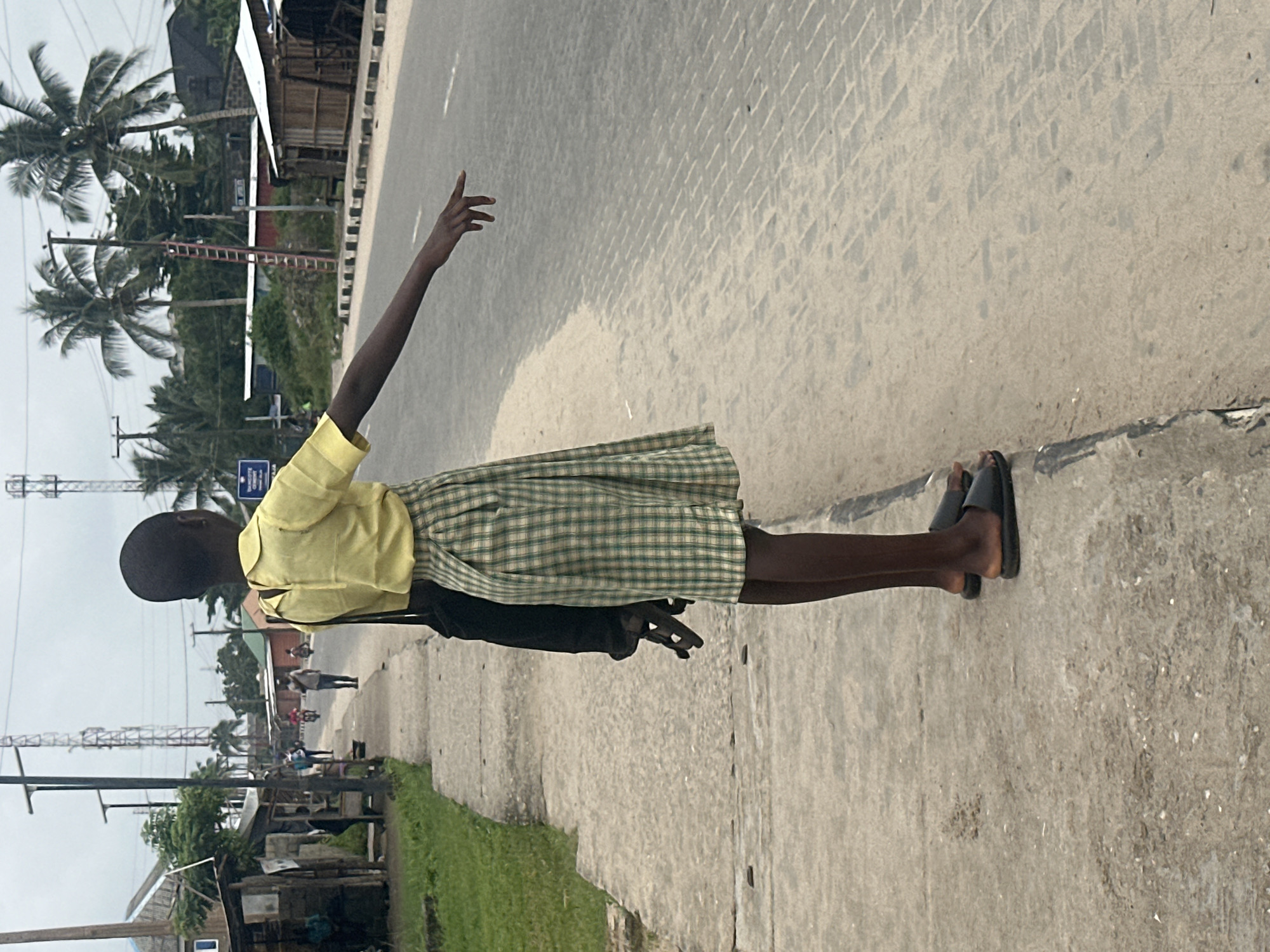 A young person, seen from the back, stands on a concrete sidewalk next to a road. They are wearing a yellow collared shirt and a green and white plaid skirt, along with black slip-on sandals. The person's right arm is extended forward, with their index finger pointing towards the road. They are carrying a black bag over their left shoulder. In the background, a paved road runs parallel to the sidewalk, with a white dashed line marking the lane. The environment appears to be a semi-urban area with buildings and vegetation visible. Communication towers are present in the distance. The scene suggests the individual may be hitchhiking or indicating a direction. This location is identified as being in Badagry, Nigeria.