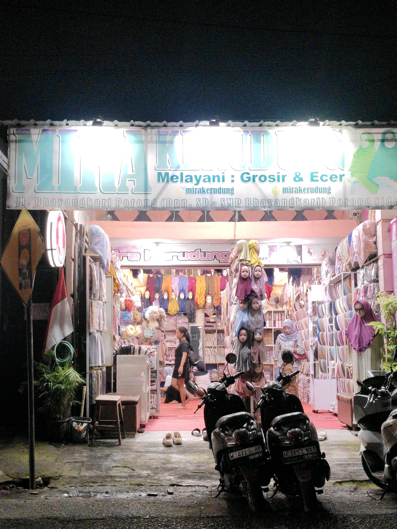 At nighttime in Juwet Kenongo, Indonesia, a storefront named "MIRAKERUDUNG" is brightly illuminated by overhead lights. The shop's facade features large, stylized lettering, and smaller text indicating it offers "Melayani : Grosir & Ecer" (Serving: Wholesale & Retail) and the business name "mirakerudung" repeated. Below this, an address is visible: "Jl. Bhayangkari Porong (Dpn. SD - SMP Bhayangkari)) Porong Sido".
Inside the shop, rows of colorful hijabs and clothing are displayed on shelves and racks. Several mannequins dressed in hijabs are placed near the front, with a younger woman in a black outfit standing to their left. Further inside, a woman in a light-colored patterned dress is visible, and another woman in a pale blue hijab is browsing items.
Outside the shop, the wet, dark asphalt of a street is visible, reflecting the light from the store. Two scooters are parked in front of the entrance. One scooter, with license plate H 4725 RP, is closer to the left, and another with license plate N 3653 NAQ is to its right. A small wooden stool and a potted plant are placed on the left side of the sidewalk, next to a yellow traffic sign and a red and white Indonesian flag. Several pairs of shoes are scattered on the ground near the shop entrance. The overall atmosphere suggests a busy retail environment open late into the evening.