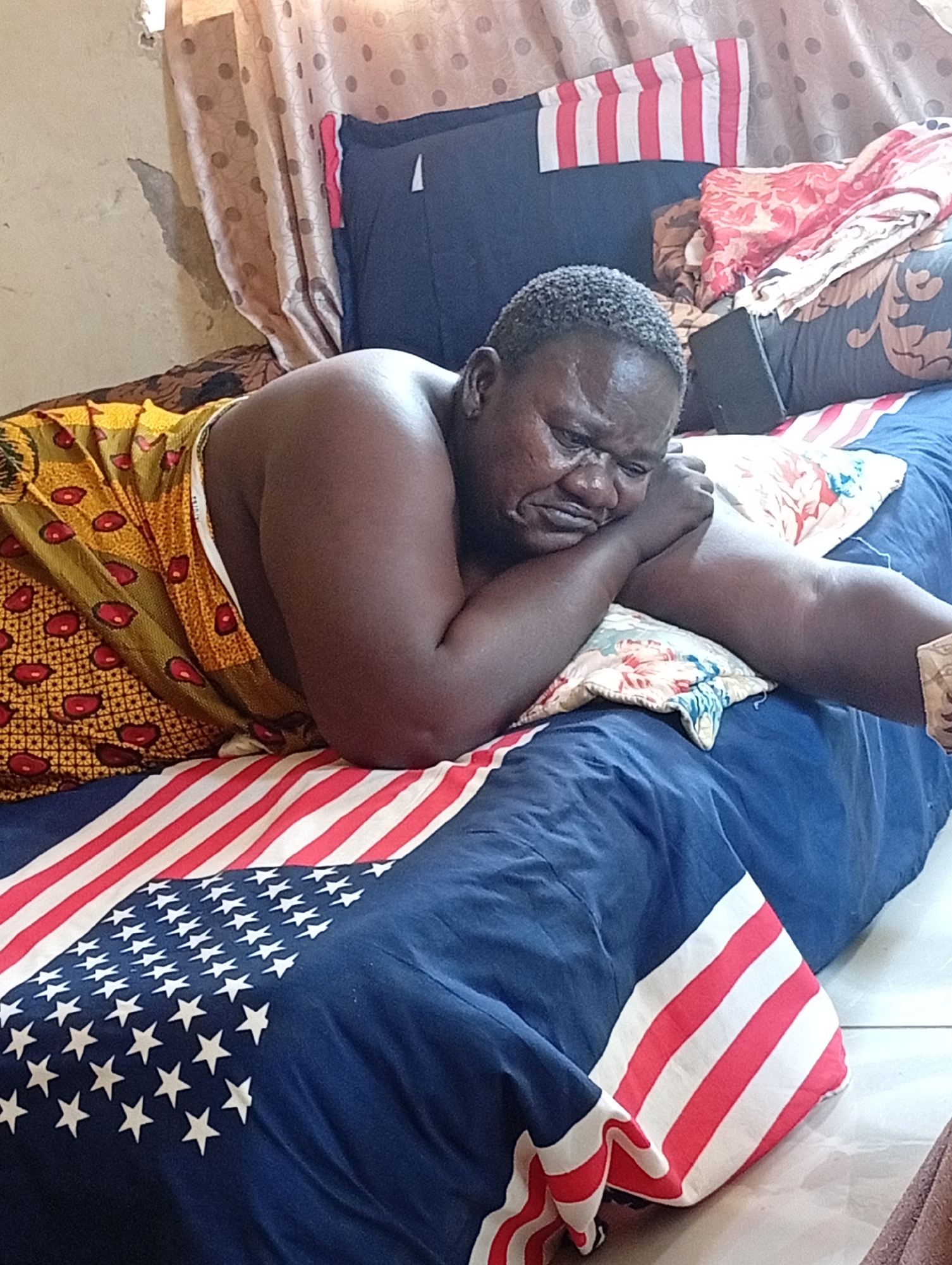 An adult individual is depicted lying on a bed indoors. The person, who appears to be male with dark skin and short, textured hair, is lying on their left side, propped up on their elbow with their head resting on their hand. Their facial expression conveys sadness, worry, or deep thought, with a downturned mouth and eyes cast downwards. The individual is bare-chested and wears a vibrant, patterned fabric, possibly a wrapper, in yellow, olive green, and red/brown designs around their lower body.
The bed is covered with blue bedding featuring a prominent American flag design, including both stars and stripes. A floral-patterned pillow supports their arm and head, and another American flag-patterned pillow is visible at the head of the bed. In the background, a light-colored wall shows signs of wear, with visible peeling paint and discoloration. Light brown curtains with a polka-dot pattern hang behind the bed. A dark, rectangular object, possibly a mobile device or book, is partially visible near the person's head, tucked into the bedding. The scene suggests a moment of quiet introspection or distress in what appears to be a modest bedroom setting in Sabon Tasha, Nigeria. No text is visible in the image.