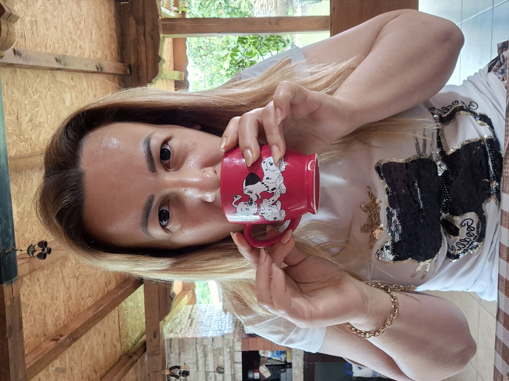 A medium shot captures a woman with blonde hair, positioned centrally and looking directly at the camera, holding a red mug with images of puppies. She is drinking from the mug, her hands cupping it. The woman is wearing a white t-shirt adorned with a large black sequined cat design and a golden crown above it. She has a gold bracelet on her left wrist. The background features a rustic, open-air structure with a wooden plank ceiling and exposed beams. To the left, a stone fireplace with decorative elements is visible. Beyond this, greenery and a window frame suggest an outdoor setting. The overall scene is casual and appears to be an outdoor seating area, possibly a patio or veranda, in a rural or suburban environment.
