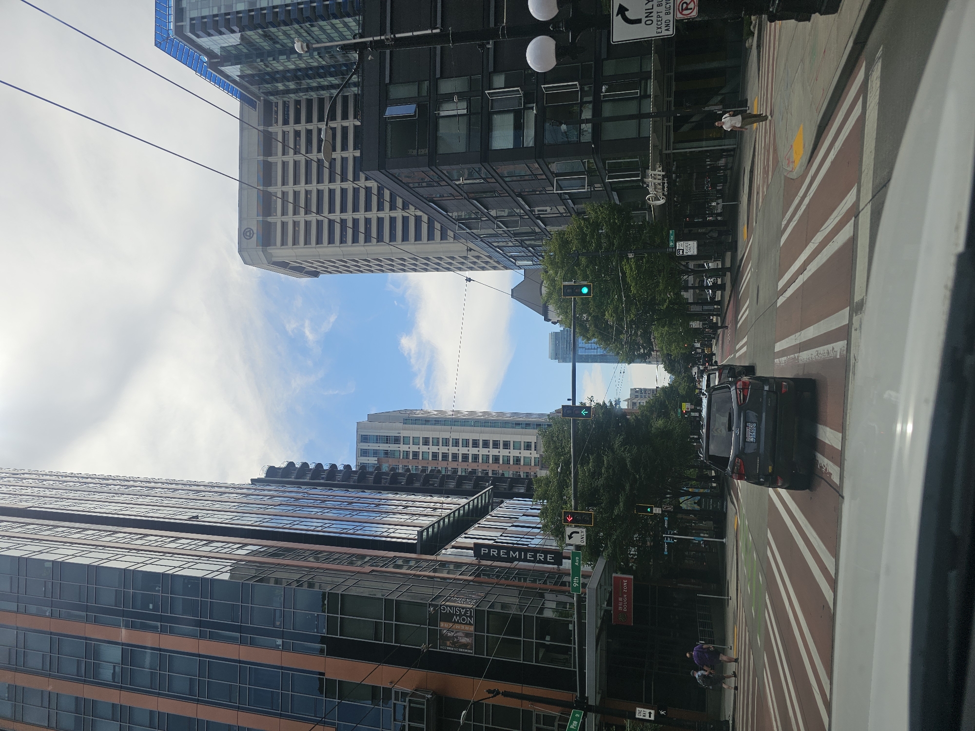 The image captures a street view in Seattle, Washington, USA, with tall buildings framing the scene. On the left, a modern building with a glass facade and vertical orange accents dominates the foreground. To the right, a darker, multi-story building with numerous open windows stands out. A black SUV is stopped at a crosswalk in the middle of the road. Pedestrians are visible on the sidewalks and crossing the street. Traffic lights are illuminated, indicating that the vehicles should proceed. Trees line the street, providing shade and greenery against the urban backdrop. The sky is partially cloudy with patches of blue. Street signs for "9th Ave" and "Pine St" are visible, helping to pinpoint the location.