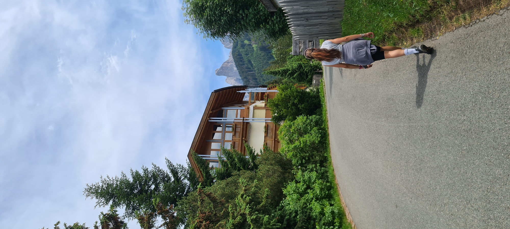 In St. Magdalena, South Tyrol, Italy, a woman walks away from the camera on a paved road. She has long brown hair, wears a grey t-shirt, black shorts, and white socks with black shoes, and has a grey jacket tied around her waist. The road is lined with green grass and a wooden fence on the right side. On the left, a multi-story wooden and glass building is visible, surrounded by lush green trees. In the background, a dramatic mountain peak rises against a partly cloudy sky.