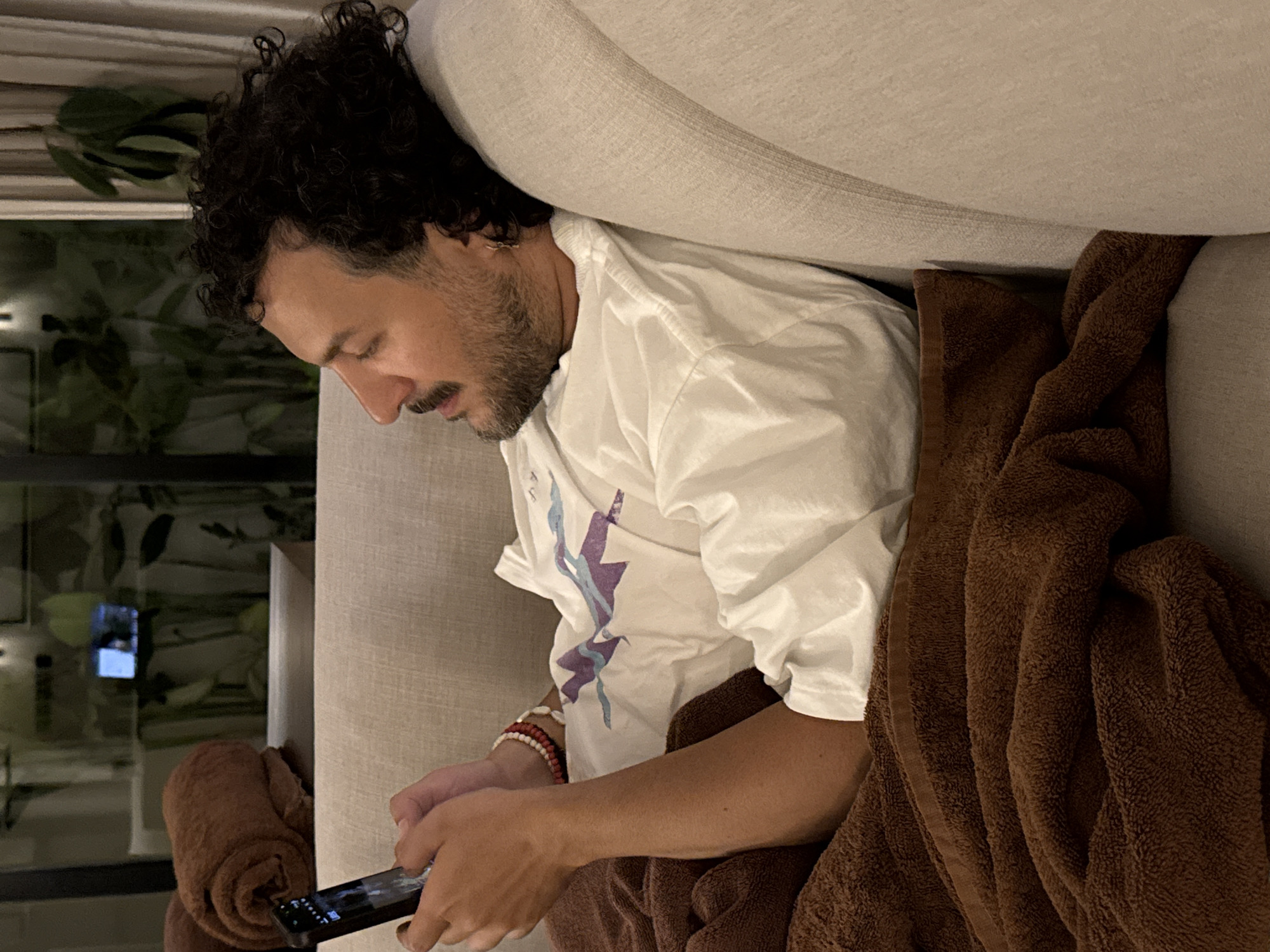 A man with dark curly hair and a beard is depicted lying on a light-colored sofa, engrossed in his smartphone. He is wearing a white short-sleeved t-shirt with a purple and light blue abstract graphic design on the chest, and several beaded bracelets on his right wrist. His gaze is directed downwards at the screen of the black smartphone he holds with both hands.
A soft, brown textured blanket is draped over his lower body and the sofa, creating a cozy atmosphere. The sofa itself is made of a light beige or off-white fabric. Behind the man, a large window or glass partition reveals a dimly lit outdoor or adjacent area with visible green foliage, suggesting evening or nighttime. Reflections on the glass include a small, bright rectangular light, possibly a screen or television, and another light fixture. To the far left, a rolled-up brown towel or blanket is visible on the sofa.
The scene suggests a moment of leisure and relaxation, likely indoors, possibly in a living room or hotel room in Kuta Selatan, given the provided context. There is no visible text in the image.