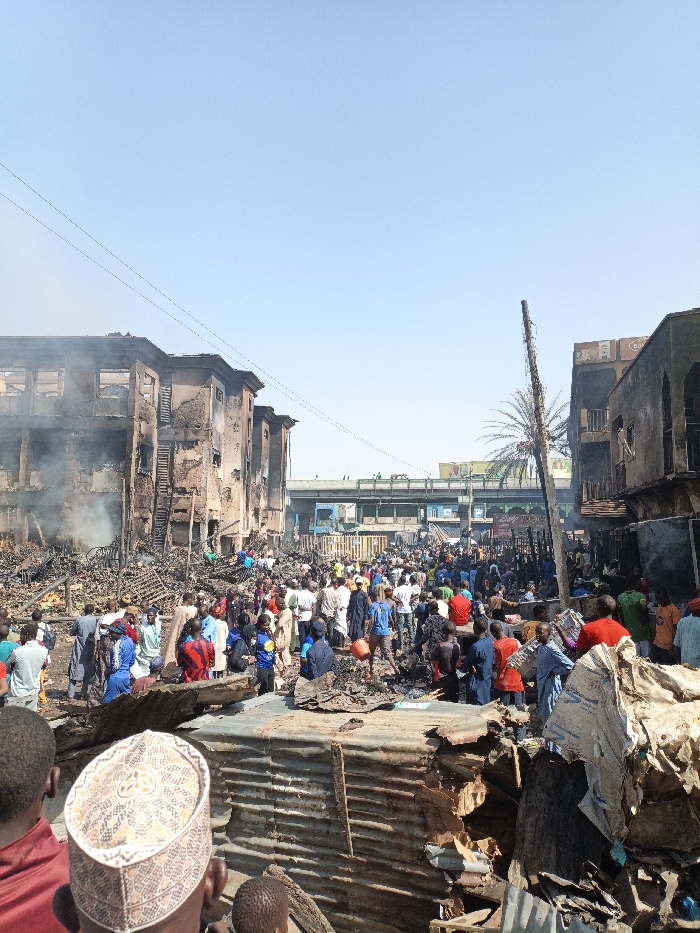 This image depicts the aftermath of a significant fire event in Fagge C, Nigeria, under a clear, bright blue sky. Extensive destruction is visible across a wide urban area. On the left, multiple multi-story buildings are severely damaged by fire, displaying blackened facades, exposed interior structures, and missing windows; faint smoke is observed rising from one structure. On the right, additional fire-damaged buildings and debris are present. The ground throughout the central and foreground portions of the scene is covered in charred rubble, twisted metal, and remnants of destroyed market stalls or smaller structures, including corrugated metal sheeting. A large crowd, estimated to be over one hundred individuals, predominantly male, occupies the damaged area, observing the scene and navigating through the debris. They are dressed in various traditional and modern clothing. In the immediate foreground, the back of a person wearing a patterned kufi cap is visible. A tall, charred wooden utility pole and a palm tree are prominent on the right side of the frame. In the background, a multi-lane concrete overpass with vehicular traffic (trucks and cars) is visible. Overhead utility wires stretch across the upper portion of the frame.