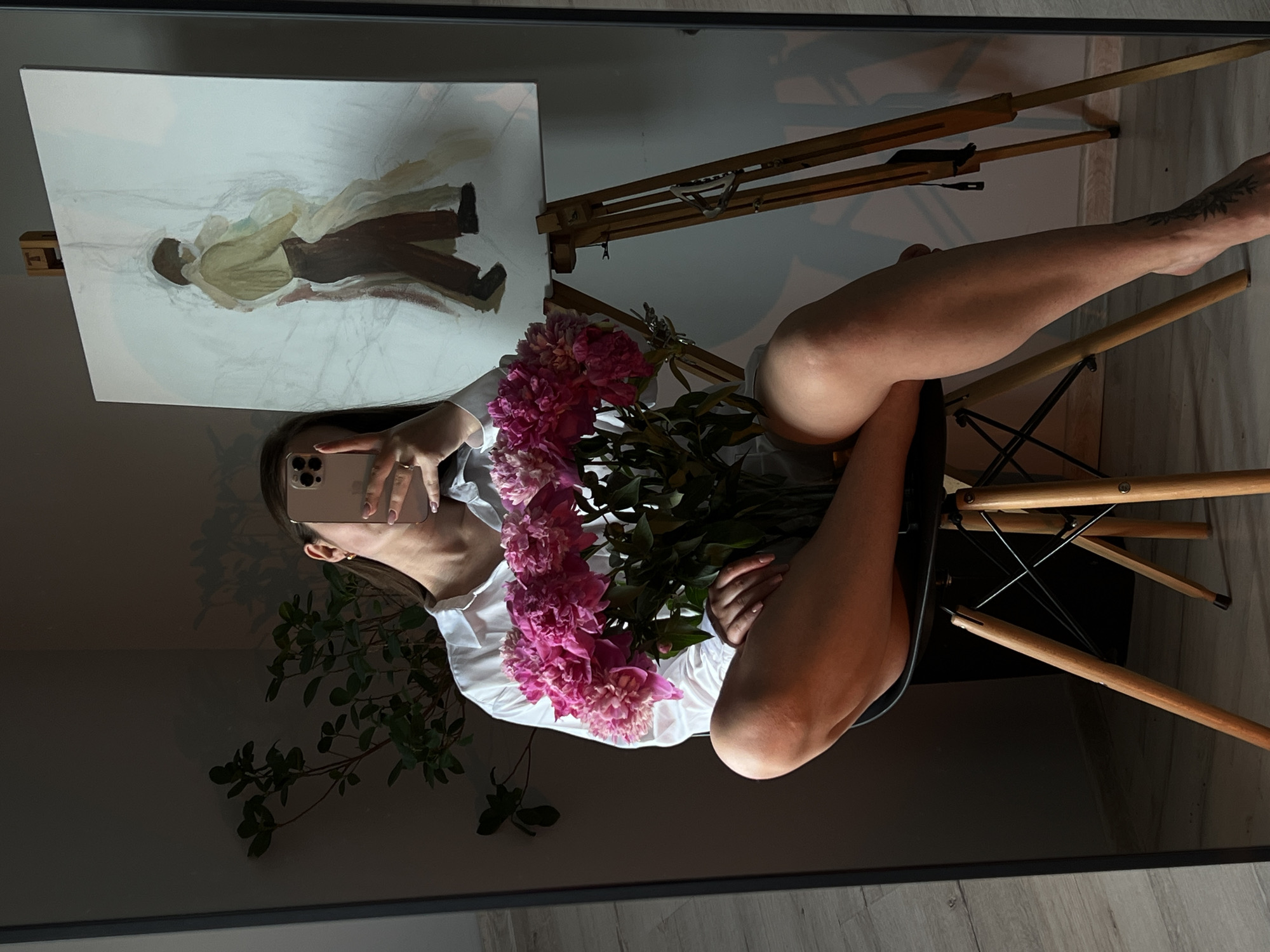 A person is sitting on a chair in front of a mirror, holding a bouquet of pink flowers and a smartphone. The person is wearing a white shirt. In the mirror's reflection, a painting of two figures can be seen on an easel. The room is sparsely decorated with a plant in the background. The lighting suggests it is daytime. The location is identified as Ternopil, Ukraine.