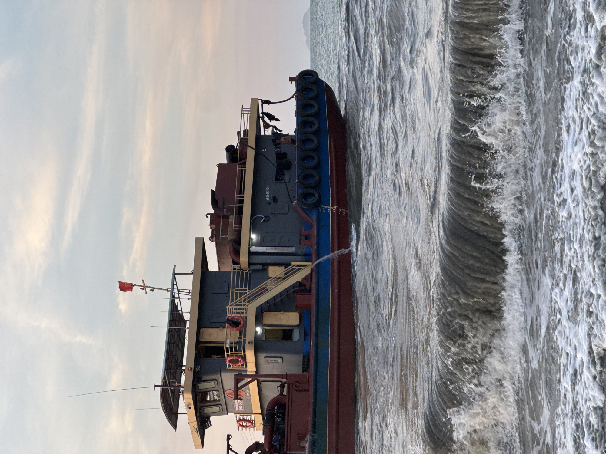 The image shows a coastal scene in Đà Nẵng, Vietnam. A large, multi-level workboat is positioned near the shore, with waves crashing against its hull. The boat has a red, blue, and grey exterior. The sky is overcast with light-colored clouds. In the background, a hazy coastline is visible.