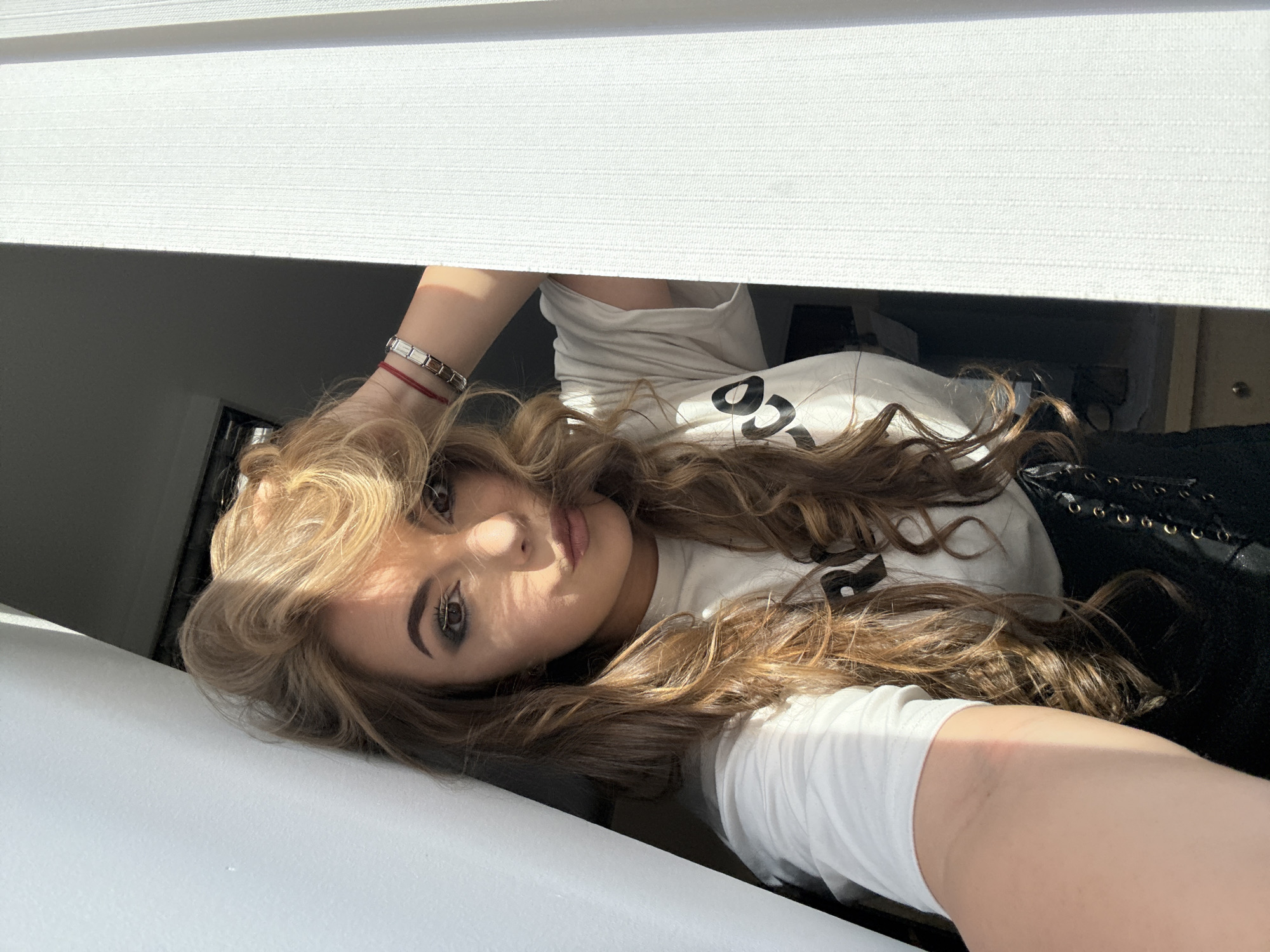 A young woman is depicted in a selfie portrait taken indoors. She has long, wavy blonde hair and is wearing a white t-shirt. She is also wearing a bracelet on her left wrist. Her eyes are accented with makeup. The photograph is taken at an unusual angle, looking upward towards the blinds or window frame above her. It is not possible to ascertain the location as Sofia, Bulgaria from this image.