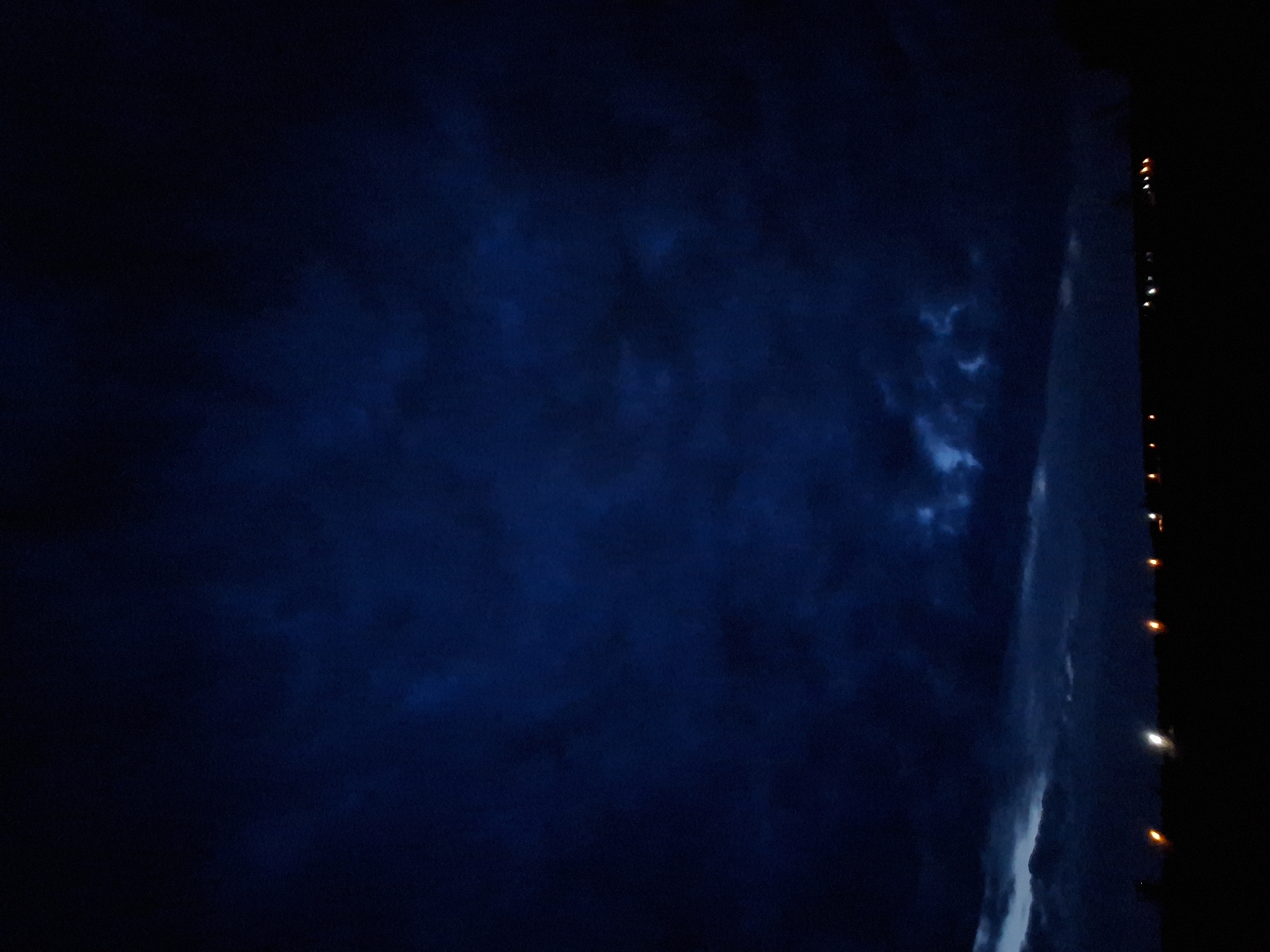 A dark, night scene in Khlevnoye, Russia. The sky is predominantly a deep, navy blue with areas of cloudy texture. To the right, a vertical structure is visible, possibly a tall building or tower, illuminated by a series of orange and white lights along its edge. The structure's surface appears to be lit by internal or reflected light, revealing some cloud-like patterns. The overall impression is one of darkness punctuated by artificial lights in an urban setting.