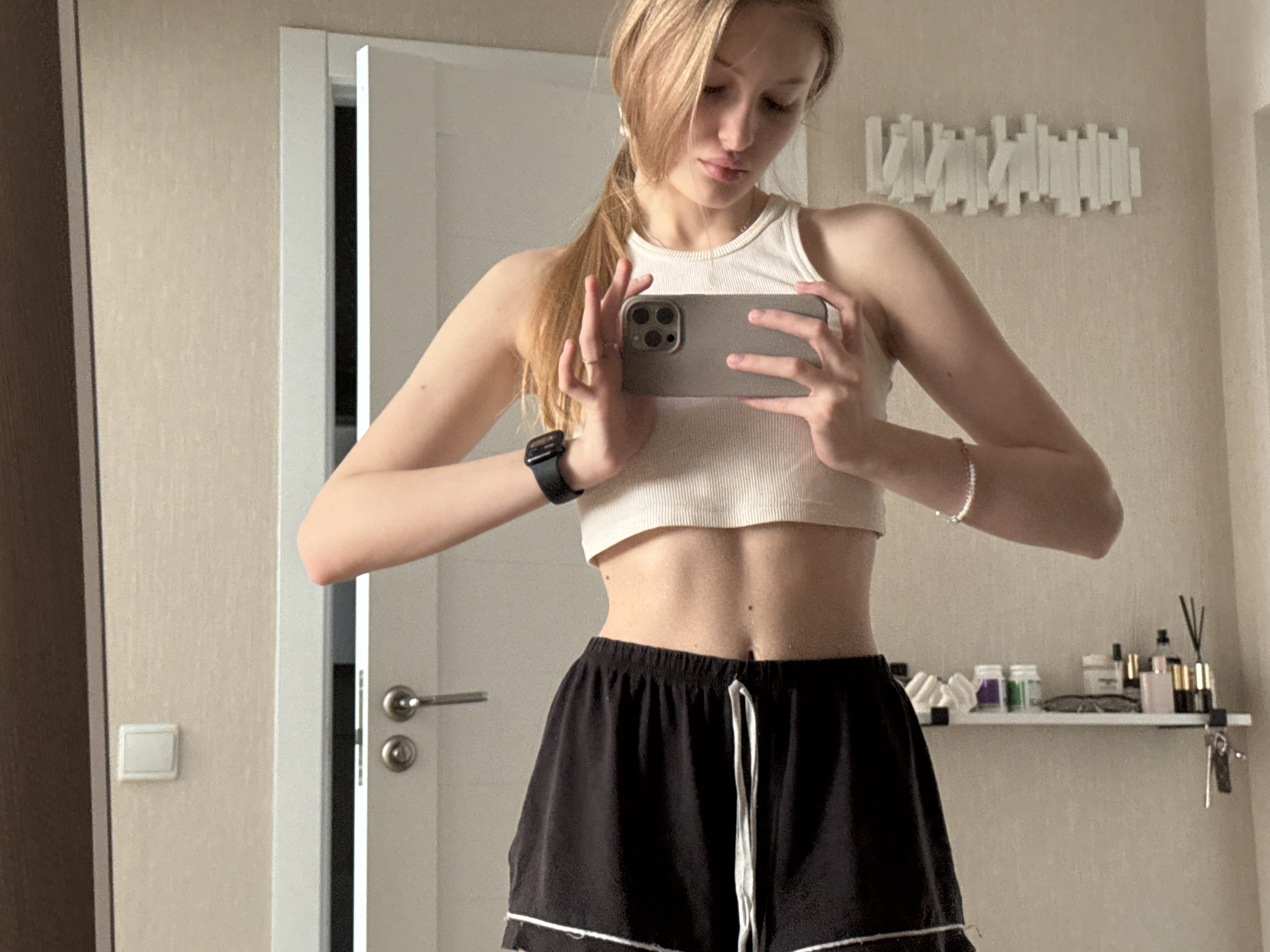 A young woman is taking a mirror selfie. She is wearing a white cropped tank top and black shorts with white trim. She is holding a gray smartphone, likely an iPhone, to take the photo. She has blonde hair pulled back in a ponytail. On her left wrist, she wears a black smartwatch and on her right a delicate bracelet. Behind her, there's a white door and a shelf with various beauty and cosmetic products. Above the shelf is a decorative white art piece. The location is possibly an apartment in Perm, Russia, based on previous user data and the interior style.
