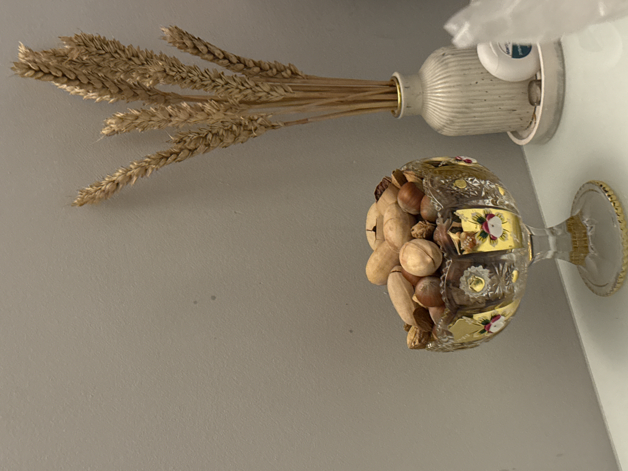 The image shows a still life composition against a pale grey wall. A bundle of dried wheat stalks is arranged in a light-colored, ridged vase, mounted on the wall. Below the vase, a decorative glass chalice filled with various nuts is positioned. The chalice has a clear glass stem and bowl with gold detailing and floral accents. The lighting suggests an indoor environment.