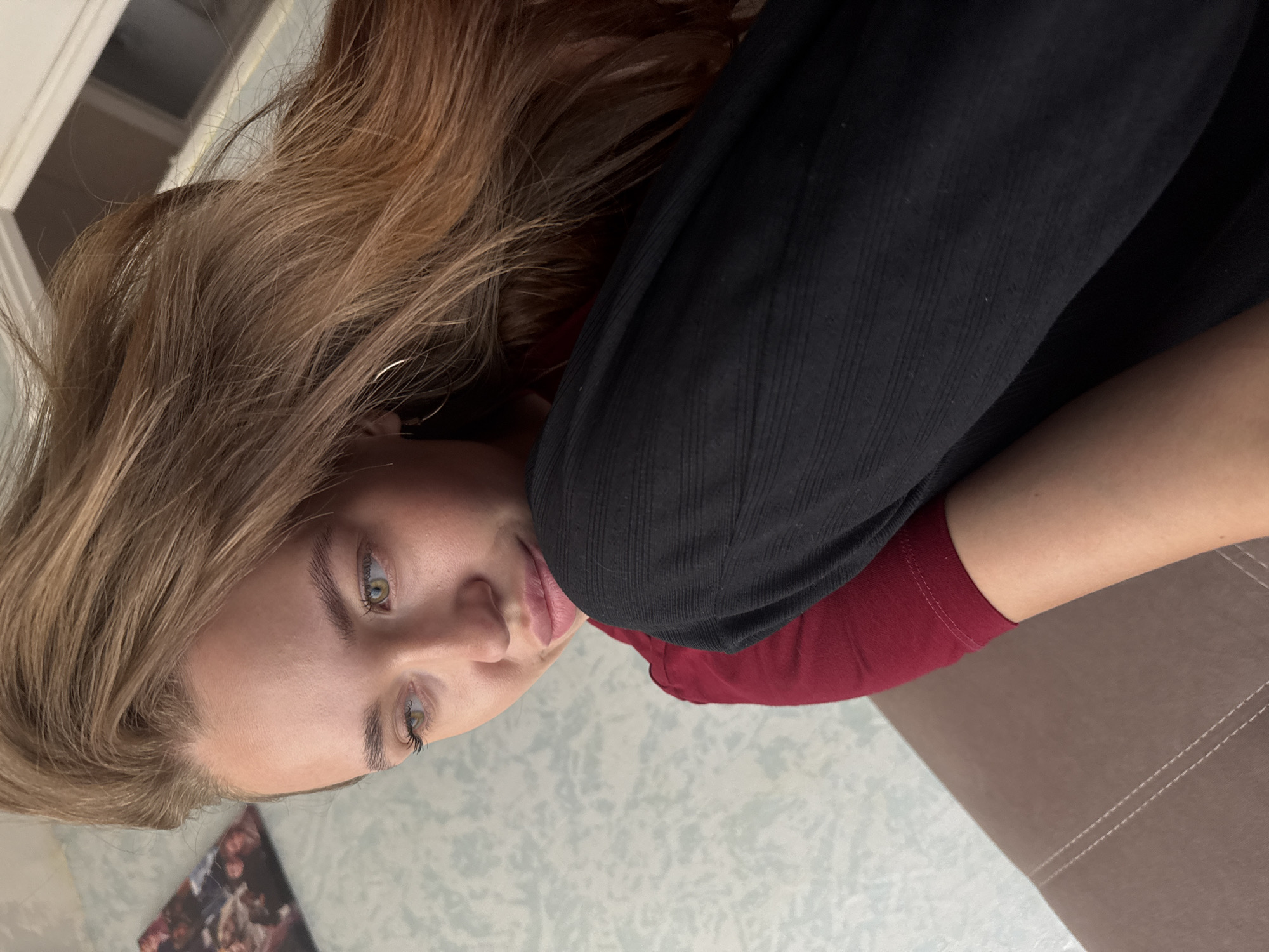 The image shows a close-up, high-angle shot of a young woman with long, wavy, light brown hair. She is wearing a dark red t-shirt and black pants. The woman is looking to her left with her eyes slightly narrowed. Her lips are slightly parted. She has a small hoop earring in her visible ear. Her left arm is bent, and her hand is resting near her face. The background is softly blurred, but a patterned wallpaper and a sofa are discernible. There is no indication of the location being in the city of Chaikovsky, Russia.