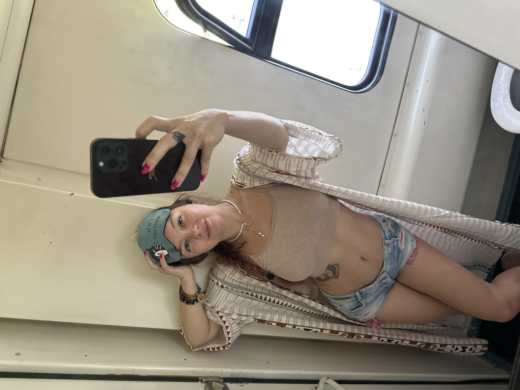 A woman is taking a selfie in a narrow, light-colored interior space, possibly a train or bus compartment, with a window visible on the right. She is wearing a beige crop top, denim shorts with distressed details and pink trim, and a patterned kimono-style robe. A teal sleep mask with a cat design is perched on her head. She is also wearing a pearl necklace and several bracelets on her left wrist. Her right hand holds a dark-colored smartphone, capturing the reflection. The walls are a neutral, off-white color, and a dark blue floor is visible at the bottom. The overall scene suggests a casual, personal moment. The location is not identifiable as Старое Перепечино, Russia.