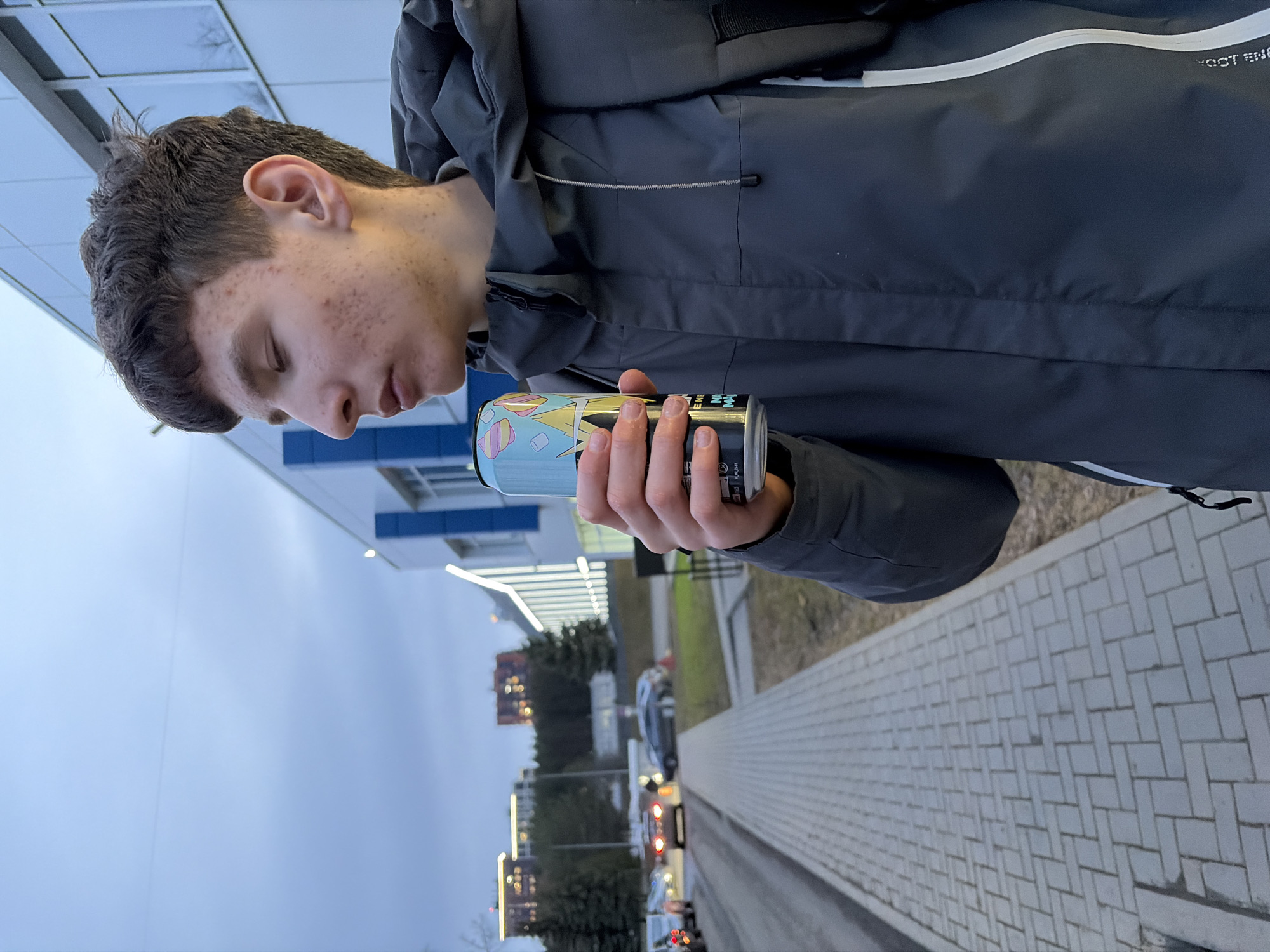 A young man is holding a can outdoors. He is wearing a gray hooded jacket. The man is looking at the can, holding it with both hands. The location is Izhevsk, Russia, as determined by reverse image search of identifiable building. The background includes a paved walkway, a building, and cars on a road. The sky is overcast.
