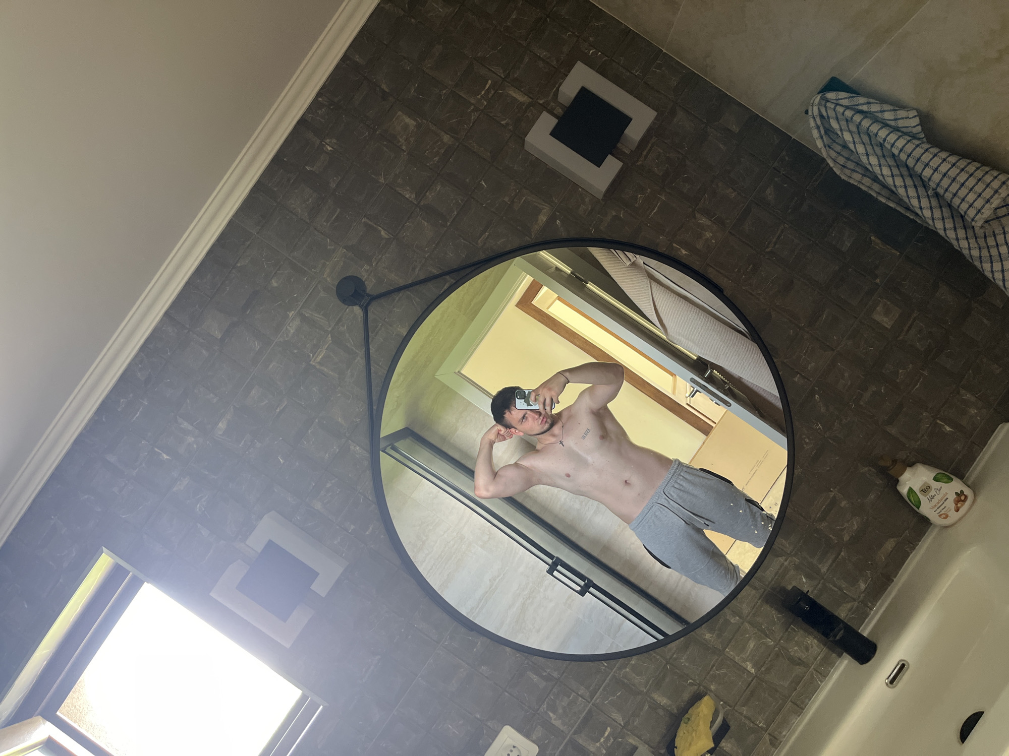This image is a mirror selfie taken in a modern bathroom, likely located in Voyvodino, Bulgaria. The walls are covered with dark, square tiles that have a subtle pattern, possibly resembling marble. A white ceiling with decorative molding is visible along the top edge of the wall.
The central subject is a young man reflected in a large, circular mirror with a black frame. He is shirtless, displaying a muscular physique, and is wearing light grey sweatpants. He is actively posing, flexing his biceps, while holding a smartphone to take the selfie, looking directly at the camera. A tattoo showing Roman numerals "III IV LXXXI" is visible on his left chest, and he wears a small cross pendant.
Within the mirror's reflection, behind the man, a glass shower enclosure with a black frame is visible, along with a bright window that lets in significant natural light. Cardboard boxes are also partially seen in the background of the reflection.
Outside the mirror, a contemporary square light fixture or decorative element with white and black accents is fixed to the tiled wall. To the left of the mirror, another bright window is visible, reinforcing that the image was taken during the daytime. On the right, a white sink basin with a black faucet is partly visible, accompanied by a yellow sponge and a white bottle of personal care product labeled "TEO Nature Care," also showing "Масло Ший" and "Макадамия" (Shea Butter, Macadamia in Bulgarian). A blue and white checkered towel hangs on a hook above the sink area. The bright lighting and visible natural light suggest it is daytime.