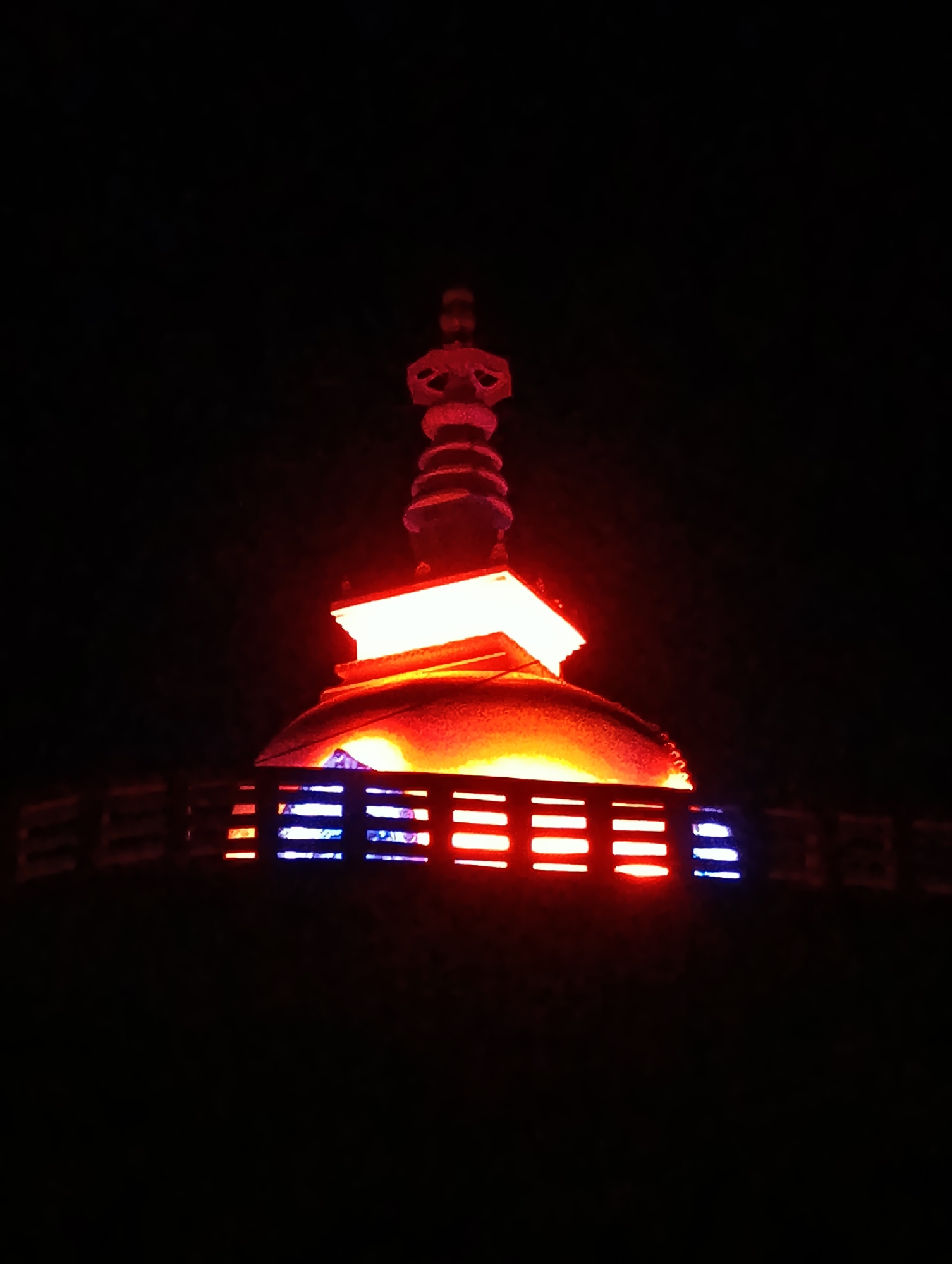 The image shows a stupa illuminated with red and blue lights at night, likely in Leh, India. The stupa, a dome-shaped structure, is the central focus. The upper portion features multiple stacked tiers, with the very top adorned with a finial. Below the dome, a flat, rectangular structure with sharp edges is visible, also lit with bright, warm light. A railing made of horizontal bars with alternating red and blue lights encircles the base of the stupa, creating a vibrant and distinct visual effect against the dark night sky. The background is entirely black, suggesting the absence of any other illuminated objects or natural light sources.