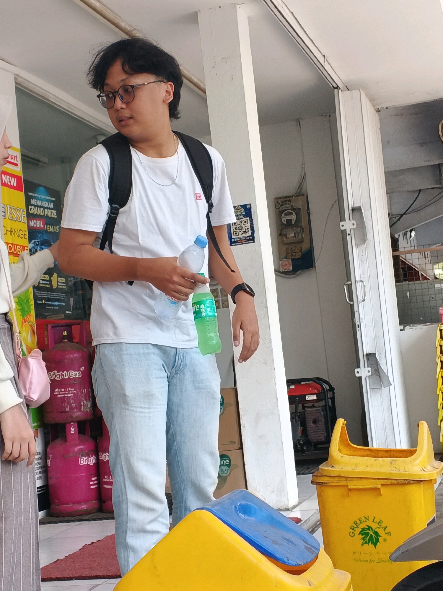 In the city of Kalirejo, Indonesia, a young man with dark, wavy hair and glasses stands outdoors, holding two bottles of drinks. He is wearing a white t-shirt, light blue jeans, and a black backpack. The man is facing towards the right side of the frame, looking away from the viewer. Beside him, a portion of another person's arm and shoulder is visible, wearing a light-colored sweater.
In the background, there are several items related to commerce and public services. To the left, a yellow and red sign is partially visible, advertising a "Grand Prize." Below the sign, two large pink gas cylinders are stacked, with one labeled "Bright Gas." Further back, a red and black generator is situated near a white wall with electrical meters and wires. In the foreground, two brightly colored trash receptacles, one yellow with a blue lid and another entirely yellow, are visible. The yellow trash can has a "Green Leaf" logo. The overall environment suggests an outdoor commercial area or a service point.