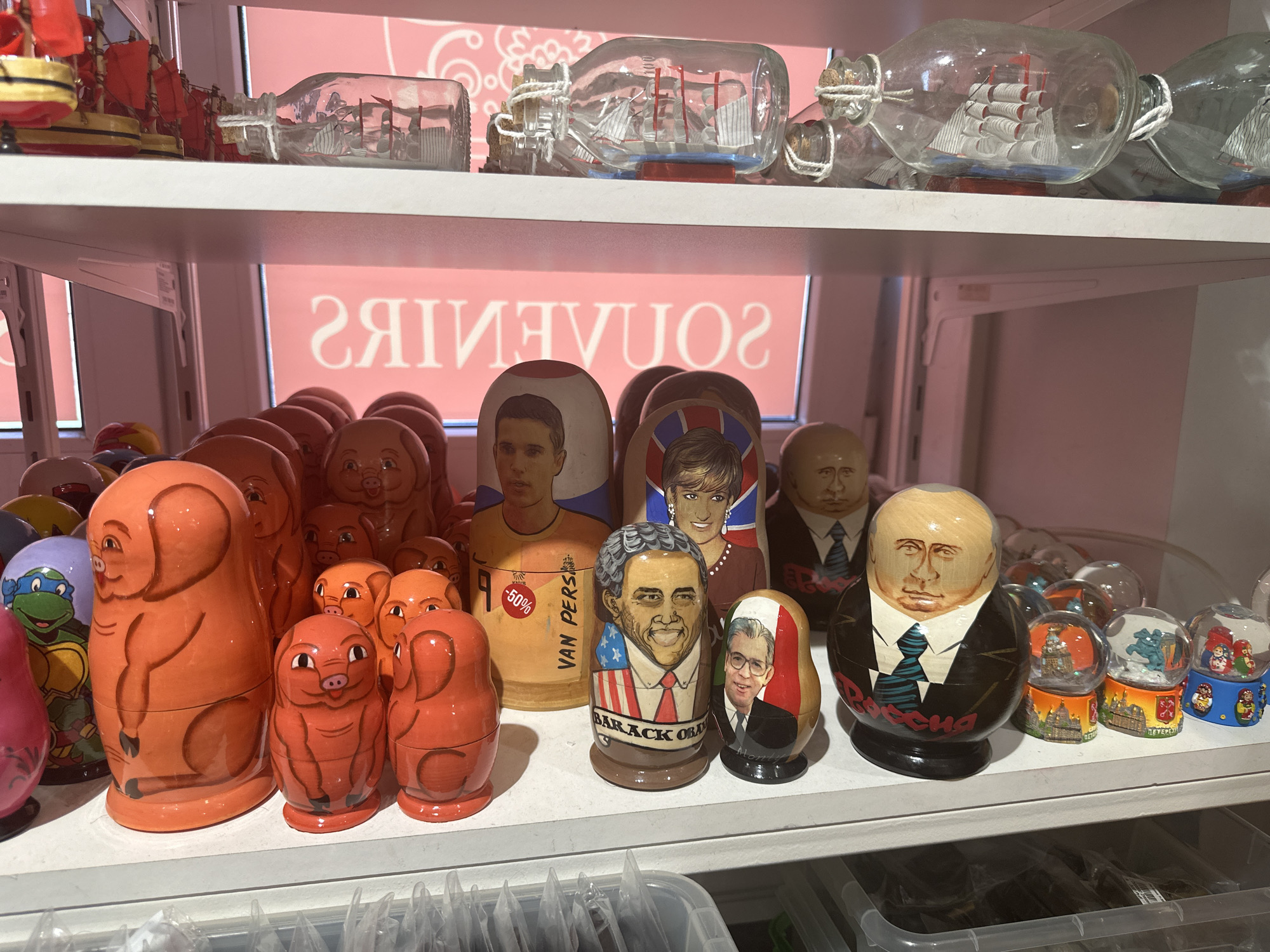 The image depicts a shelf in a shop in Saint Petersburg, Russia, displaying various souvenir items. The shelf is stocked with a collection of matryoshka dolls, also known as Russian nesting dolls, featuring caricatures of political figures such as Barack Obama, Vladimir Putin, and others. There are also matryoshka dolls with designs of animals, such as pigs and turtles, as well as more traditional Russian figures. On the shelf above, there are glass bottles containing model ships. The background features a pink wall with the word "SOUVENIRS" printed on it. The shop seems to cater to tourists seeking mementos of their visit to Russia, with the dolls and model ships being typical souvenirs.