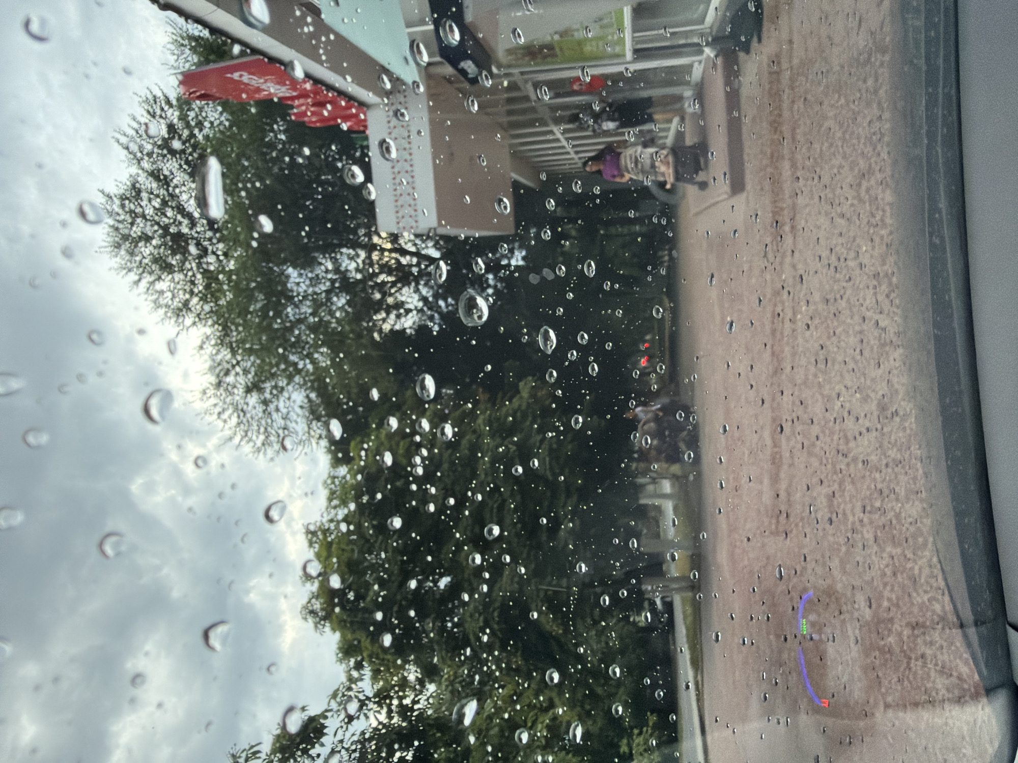 The image shows a street scene in Strășeni, Moldova, viewed through a rain-streaked windshield. The sky is overcast and cloudy. Trees line the background, and to the right, a building with a red and white sign is visible. In front of the building, a person is pushing a baby stroller. Another person can be seen further in the background. The ground is paved with reddish-brown bricks, and puddles are present from the rain. A projection on the lower left of the windshield displays a curved bar with colored segments, likely a Heads-Up Display (HUD) indicator.