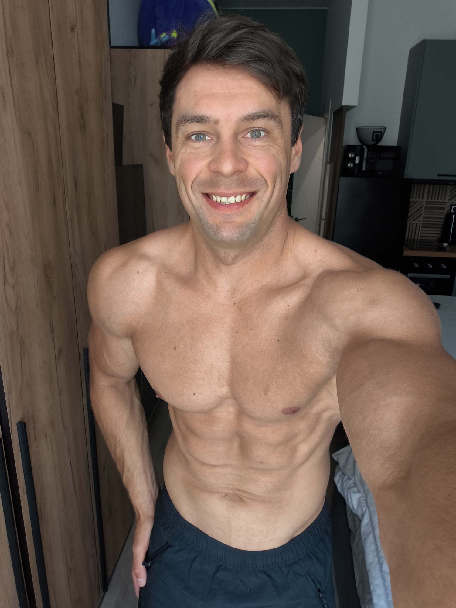 In Moscow, Russia, a man is captured in a selfie taken indoors. He is shirtless, showcasing his muscular physique with visible abs and well-defined arms. He has dark hair and green eyes, and he is smiling broadly. He is wearing dark blue athletic shorts. The background suggests a home environment, with a wooden wardrobe on the left and kitchen cabinets and appliances visible on the right. The lighting appears to be natural and from an overhead source. The overall impression is that of a personal, candid moment captured in a domestic setting.