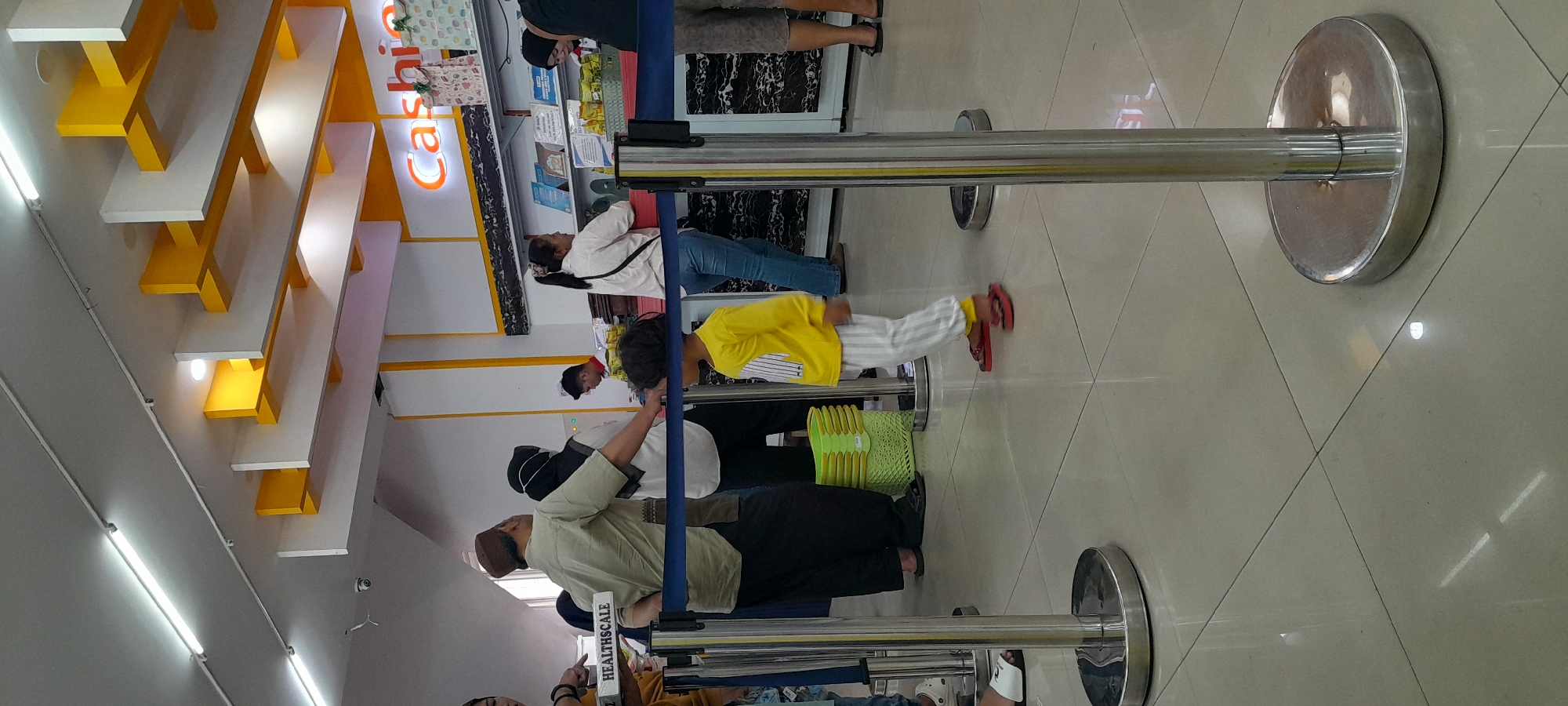 The image depicts a line of people indoors, likely a retail or service establishment in Rogojampi, Indonesia. The ceiling features modern yellow and white architectural elements with fluorescent lighting. Several individuals are standing, some in a queue, and one child in a yellow shirt and striped pants is walking. A sign reading "HEALTHSCALE" is visible on a box held by one of the individuals. Barriers with chrome stanchions and blue ropes guide the flow of people. In the background, a "Cashier" sign is illuminated, and there are shelves with products behind a counter. The floor is tiled with a light-colored, glossy finish. The overall impression is a well-lit, organized indoor space.