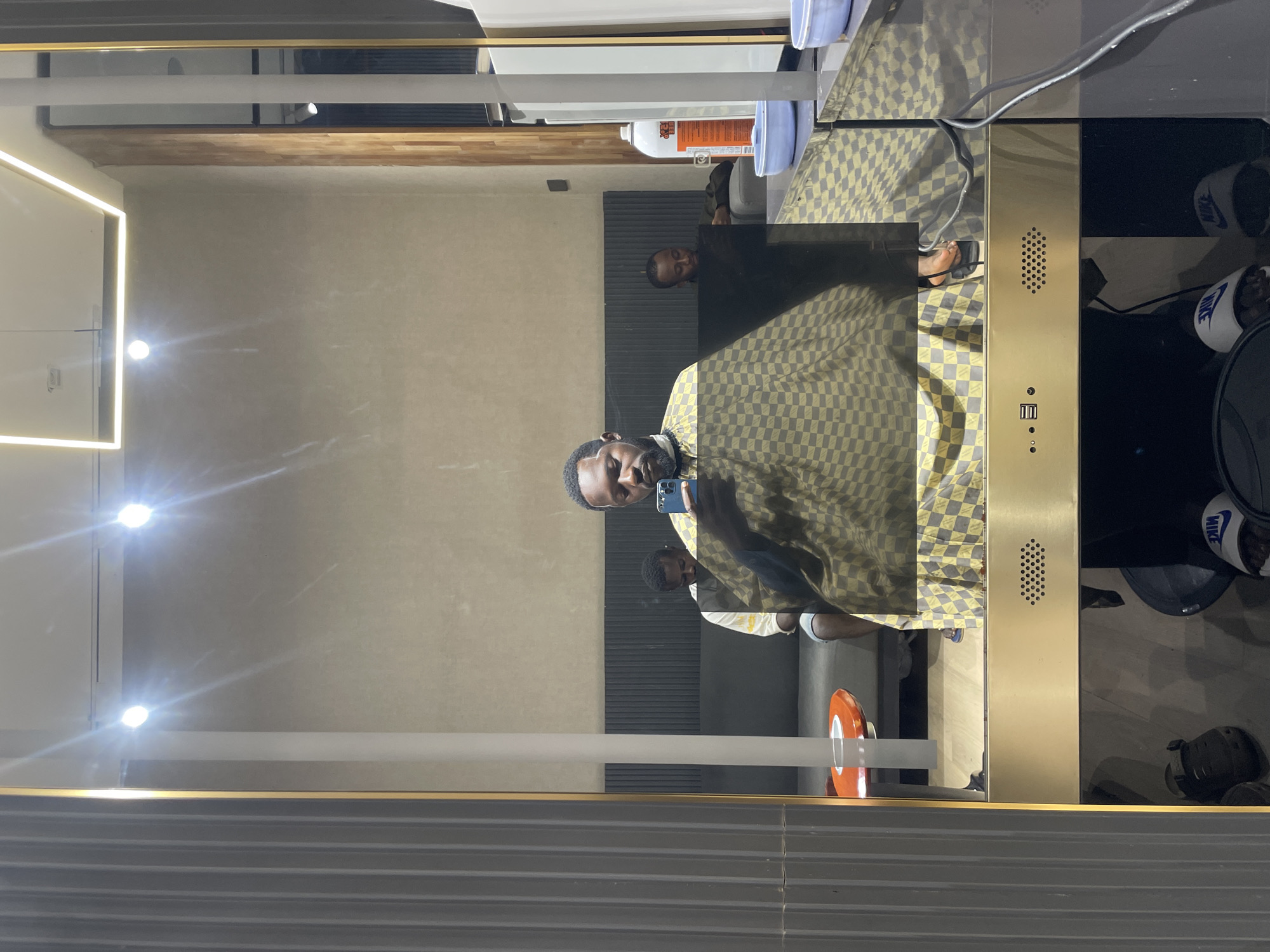 A self-portrait taken in a mirror shows a Black man in a barber's cape, holding a smartphone. He appears to be in a barber shop, with other customers visible in the background reflected in the mirror. The barber shop has modern lighting and decor. The man is wearing a black shirt, and a patterned cape. In the reflection, there are two other men seated in chairs, and a bottle of spray in the background. The floor is wooden, and there are multiple light fixtures on the ceiling. The overall impression is of a clean and contemporary barbershop environment in Guzape, Nigeria.