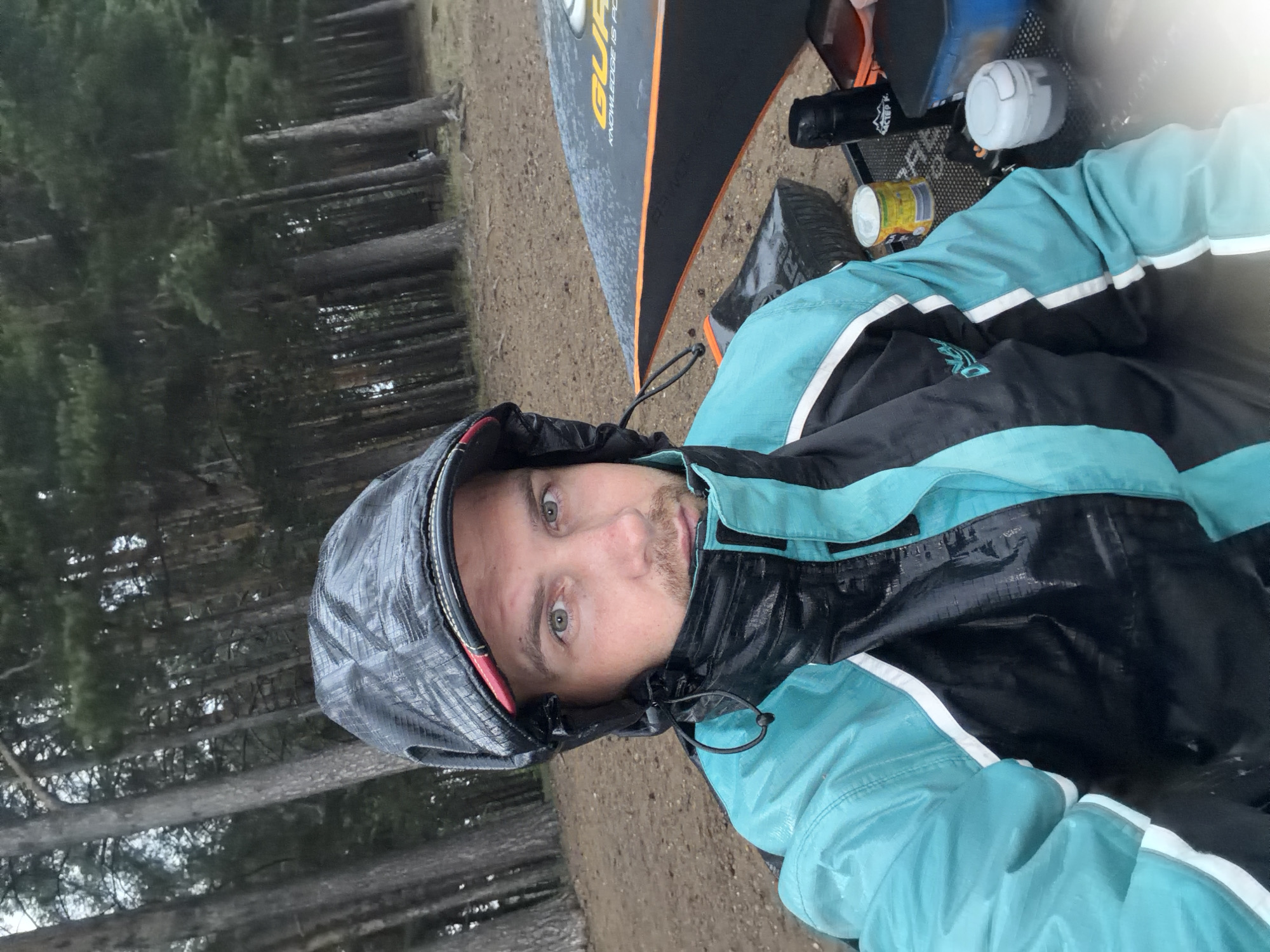 In Chaikovsky, Russia, a man is pictured in a selfie taken outdoors. He is wearing a black and turquoise hooded jacket and a black hood. The man is looking directly at the camera with a neutral expression. Behind him, a dense forest of tall, thin trees with green foliage is visible. The ground appears to be sandy and covered with fallen pine needles. To the right of the man, a black and orange umbrella is partially visible, with the word "GUR" printed on it. A black thermos, a yellow can, and a white lid are on a table in front of the man. The overall scene suggests an outdoor activity in a wooded area, possibly camping or fishing, during overcast weather.
