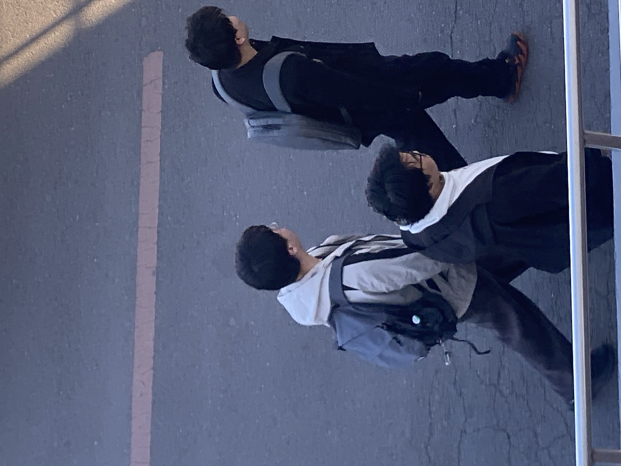 The image shows three people walking on a paved surface marked with a faded orange line, possibly a sidewalk or pathway. The top person is wearing dark clothing and a backpack. The middle person is wearing glasses and a black and white jacket. The bottom person is wearing a light-colored hooded sweatshirt and a backpack. The people appear to be walking in the same direction. A railing is present on the right side of the image. The location is possibly Zhongtun, China.