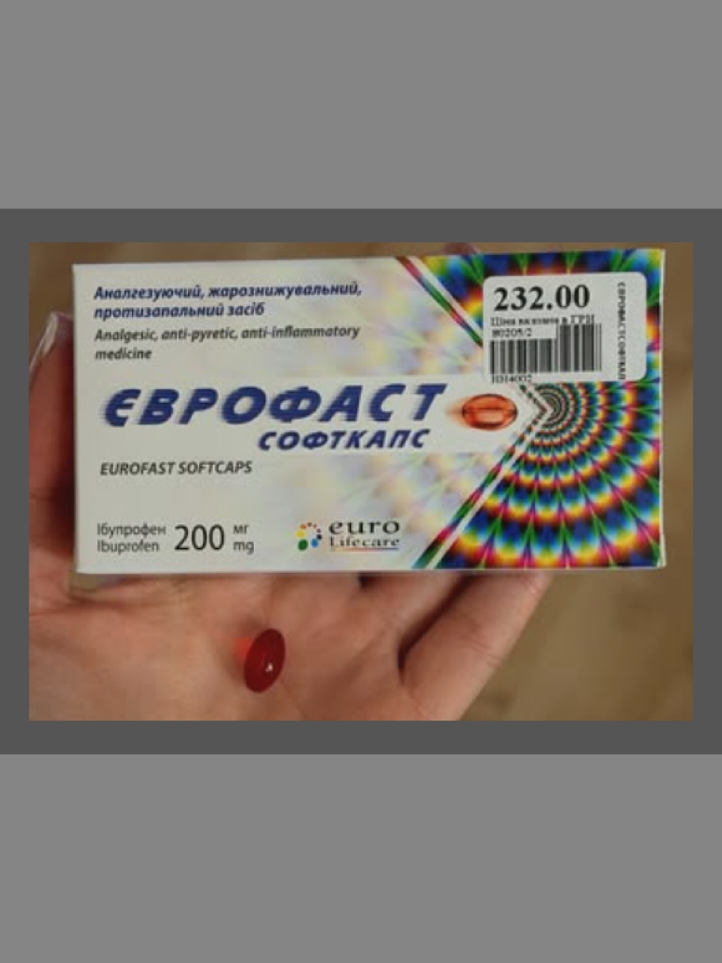 A hand holds a red pill in front of a box of "Eurofast Softcaps". The box is mostly visible, displaying text in Ukrainian and English. Text on the box reads "Анальгезуючий, жарознижувальний, протизапальний засіб" and "Analgesic, anti-pyretic, anti-inflammatory medicine". Below this, the brand name "ЄВРОФАСТ" and "EUROFAST SOFTCAPS" are prominently displayed. The pill's dosage is indicated as "16упрофен 200 мг" and "Ibuprofen 200 mg". A price tag of "232.00" is visible on the top right of the box. The background appears to be a neutral, indoor setting with the focus on the product.
