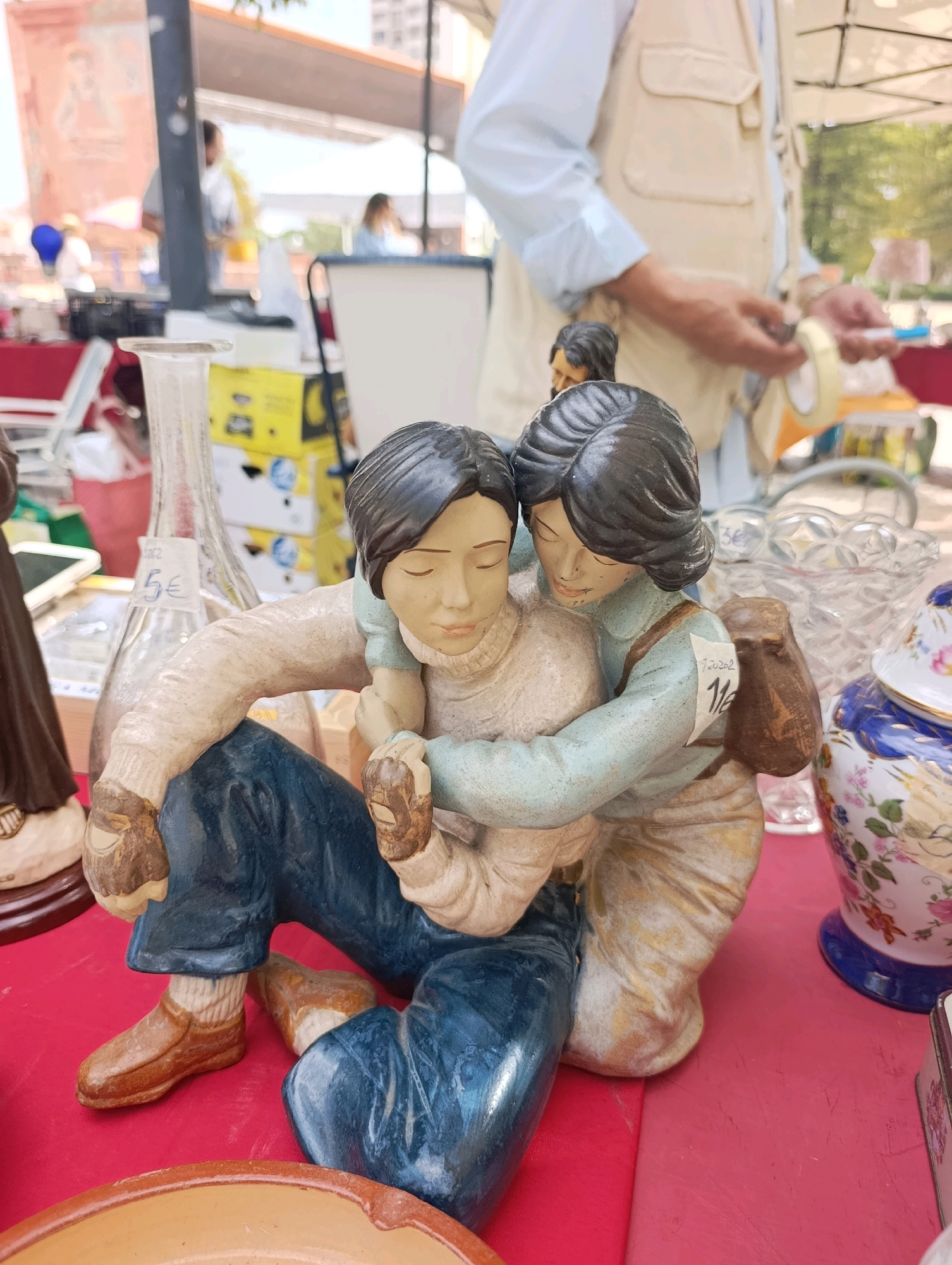 An outdoor market stall in Setúbal, Portugal displays various items, including a ceramic figurine of two people embracing. The figurine, positioned in the foreground on a red surface, depicts one figure with dark hair and a light blue shirt, embracing another figure wearing a cream-colored sweater and blue pants. Both figures have closed eyes. Behind them, a glass bottle with a price tag of "5€" and a clear glass bowl are visible. Further back, a man wearing a light vest over a blue shirt is holding a roll of tape. Other market stalls and tents are present in the background, suggesting a bustling environment. The lighting indicates a daytime setting.
