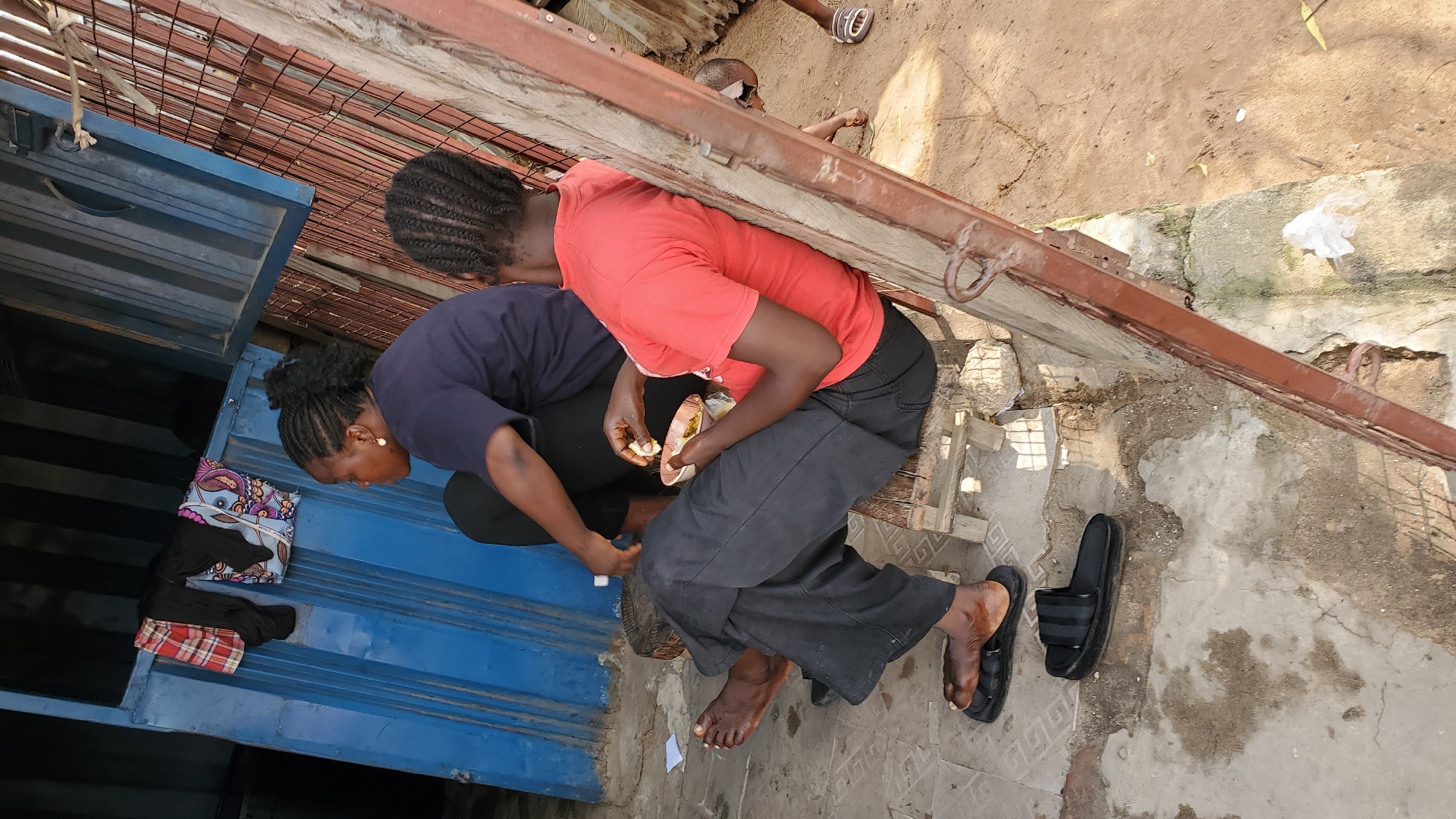 The image depicts an outdoor scene, likely in Jalingo, Nigeria, where two women are seated on a wooden bench. The woman on the left, wearing a dark blue t-shirt and black pants, is facing to her left, with her hair in braids. The woman on the right, dressed in a red t-shirt and black pants, is holding a small bowl and appears to be eating. Her hair is also braided. Both women are wearing sandals. They are positioned in front of a blue corrugated metal structure, possibly a shipping container, with some clothing hanging on it. To the right of the women, there is a rusty metal gate or structure. The ground appears to be a mix of concrete and dirt. The overall environment suggests a casual outdoor setting, possibly a market or roadside stall.