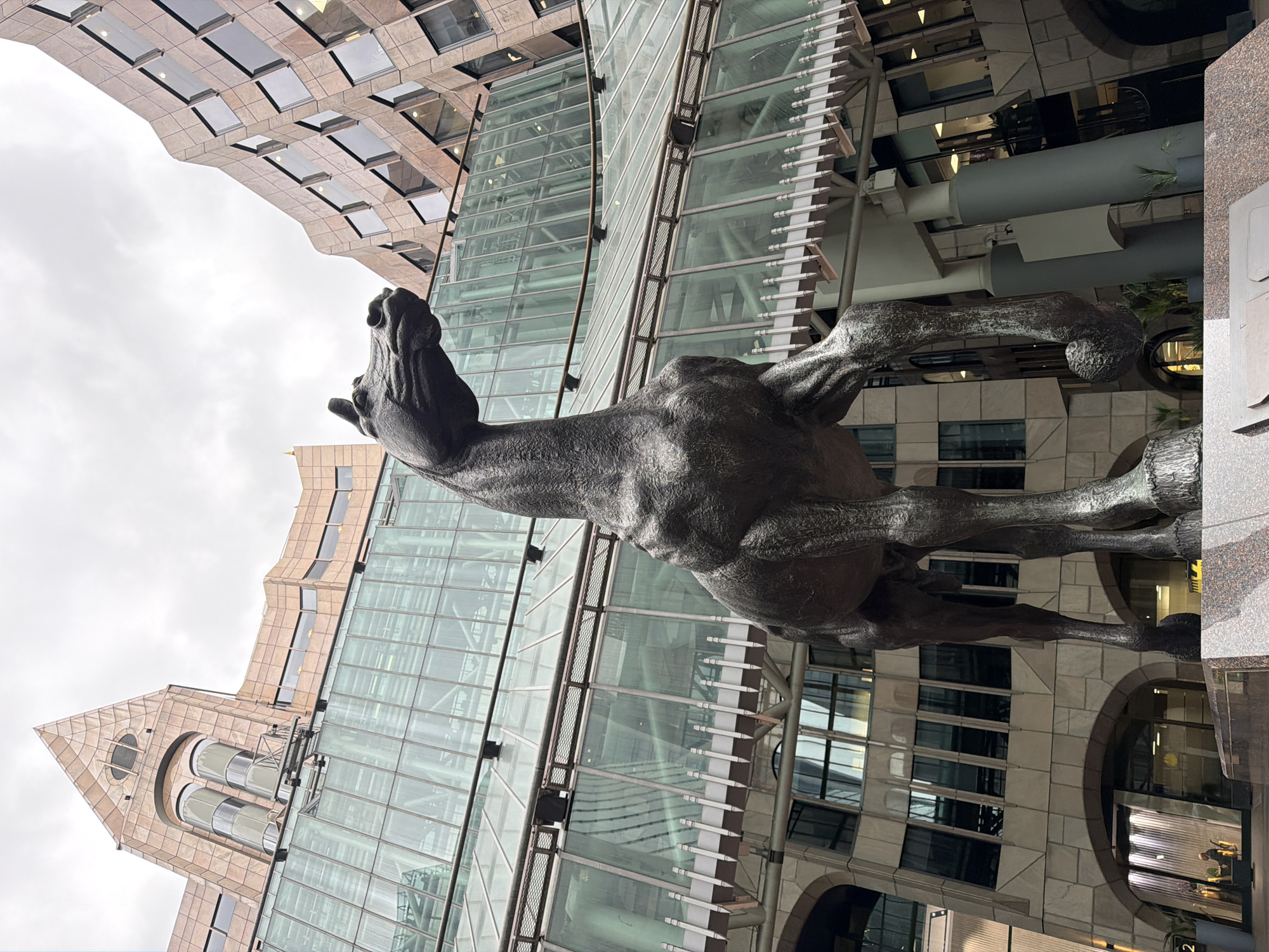 A bronze sculpture of a horse rearing up, seen from a low angle, is the central focus. The sculpture is positioned against the backdrop of a modern cityscape. The buildings feature a mix of architectural styles, including glass facades and traditional stone exteriors. The sky is overcast. The location is the City of London, United Kingdom, specifically near Broadgate. No people are visible.