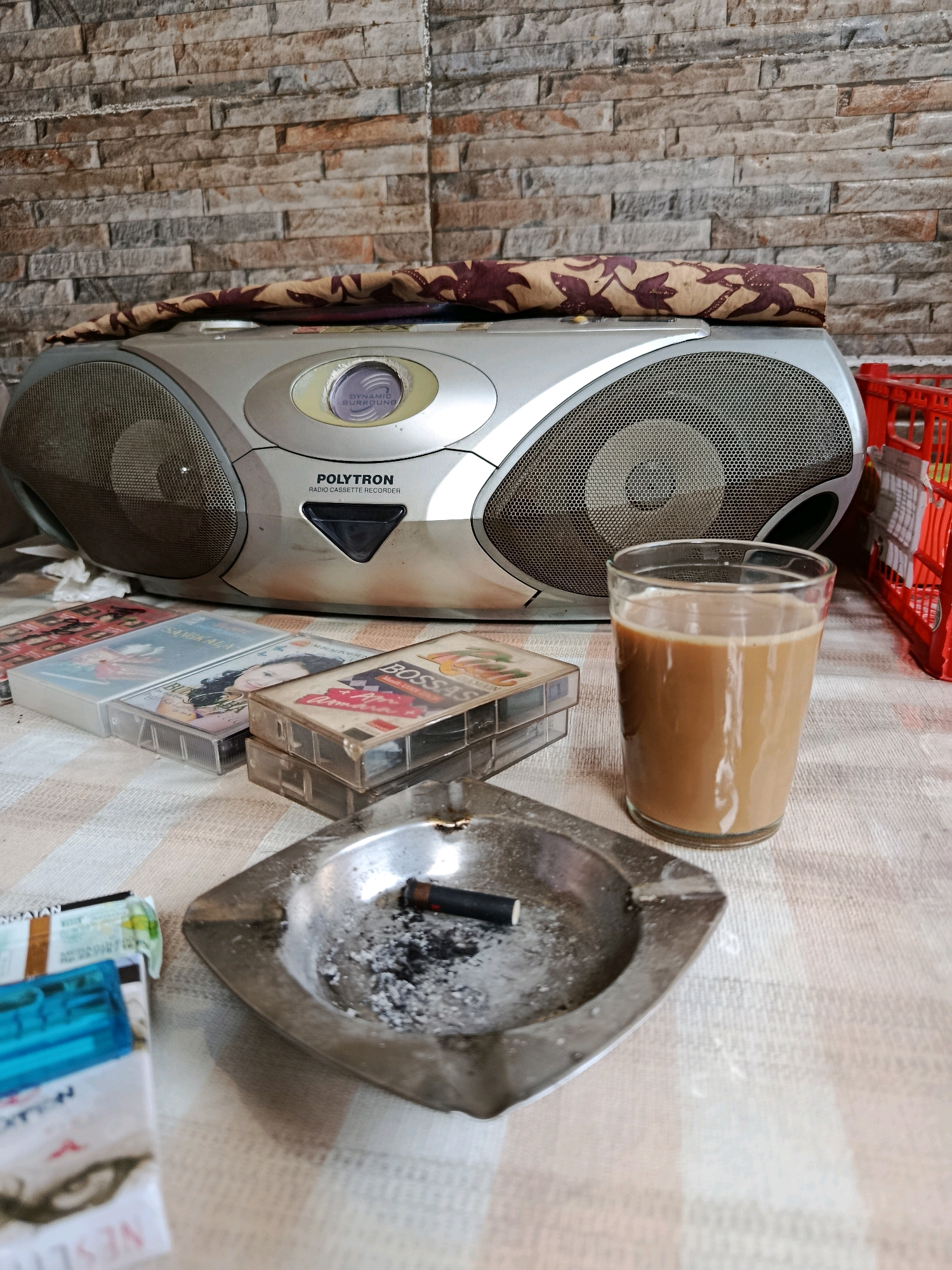 An indoor scene depicts a cluttered table with various objects. In the center, a silver Polytron radio cassette recorder is prominent, topped with a rolled-up cloth featuring a floral pattern. To the left of the recorder are several cassette tapes, and to the right, a glass of light brown liquid, likely coffee or tea, is placed. In the foreground, an ashtray contains a cigarette butt and ash, with cigarette packs nearby. The background features a textured brick wall. The overall impression is one of a casual, perhaps slightly messy, personal space. The scene is suggestive of a setting within a residential area in Bekasi, Indonesia, given the common availability of such items and the typical home decor.