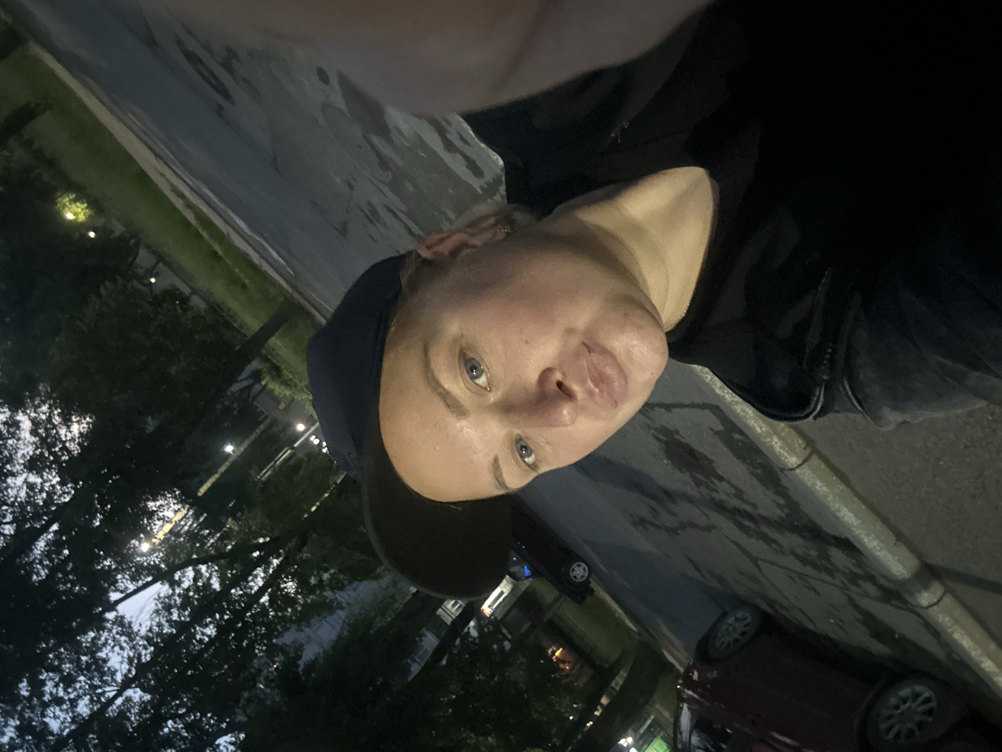 A selfie is taken by a woman in a black baseball cap and black t-shirt. She is outdoors at night, with trees and streetlights visible in the background. The street is wet, suggesting it has recently rained. Cars are parked on the side of the road. The woman has blonde hair and is looking at the camera with her lips pursed. The location is the city of Chaikovsky, Russia.