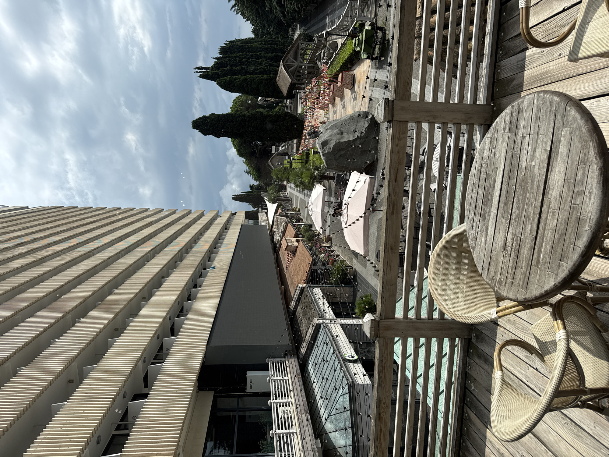 The image shows a high-angle view of an outdoor seating area on a wooden deck, with a table and two chairs in the foreground. Beyond the deck, there is a street lined with shops and trees. The scene appears to be part of a resort or tourist area. The architecture of the buildings suggests a modern design with balconies and large windows. The sky is overcast, and the lighting is diffused. There are no people visible in the immediate foreground, but there are some figures further in the distance, near the shops. The overall atmosphere is one of a relaxed, outdoor environment, likely in a warm climate.