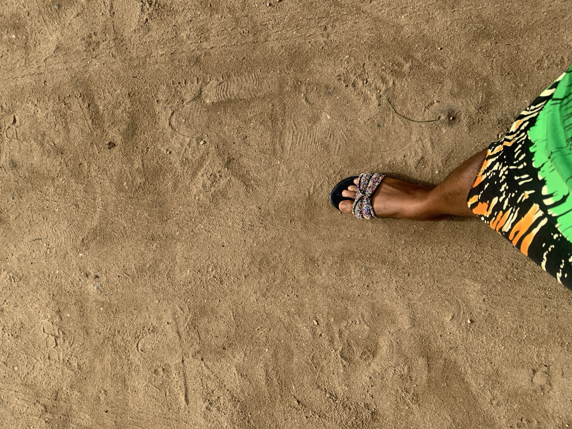 An overhead shot shows a person's leg and foot in a beaded sandal resting on a sandy, light brown surface. The person is wearing a green, black, and orange patterned garment. The sand appears to be compacted and has footprints. A small piece of straw is visible on the sand. The location is inferred to be Aba, Nigeria.