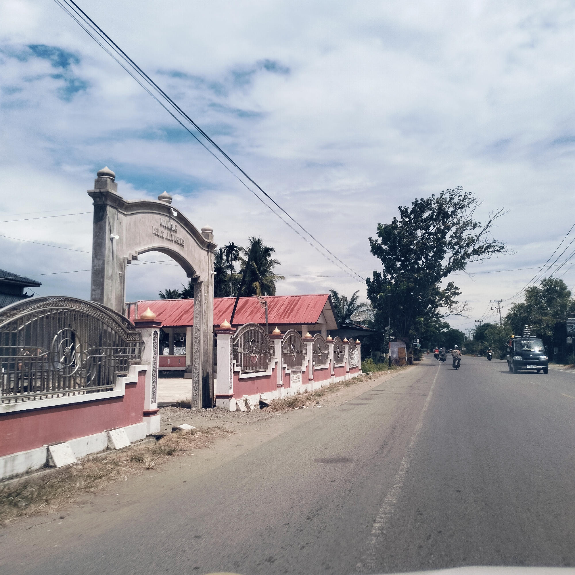 This daytime image captures a road scene in Keudealuerheung, Indonesia, under a sky with scattered clouds. The photograph appears to be taken from inside a vehicle, looking forward.
On the left side of the road, a substantial concrete archway serves as an entrance, featuring decorative elements like small dome-shaped finials and embedded lights. Inscribed on the archway is the text "MEUNSAH KEUDE ALUEREUNG". Adjacent to the archway, a decorative fence with ornate metalwork atop a red-painted lower wall extends along the roadside. Behind this fence and archway, a building with a distinctive red corrugated metal roof is visible, partially obscured by palm trees.
The asphalt road, which appears dry, stretches into the distance, showing faint white road markings in the foreground. Traffic is present on the road; a dark-colored pickup truck or van is driving towards the viewer on the right side of the road, while several motorcycles or scooters are further down the road, traveling away from the camera.
The environment is characterized by lush greenery, with numerous trees lining both sides of the road, including the prominent palm trees near the building and a large, leafy tree with some dead branches on the right. Utility poles and electrical wires are visible across the top of the frame and along the roadside, indicating developed infrastructure. The lighting suggests it is late morning or early afternoon, with clear visibility. The scene depicts an ordinary moment of daily life and transit in the area.