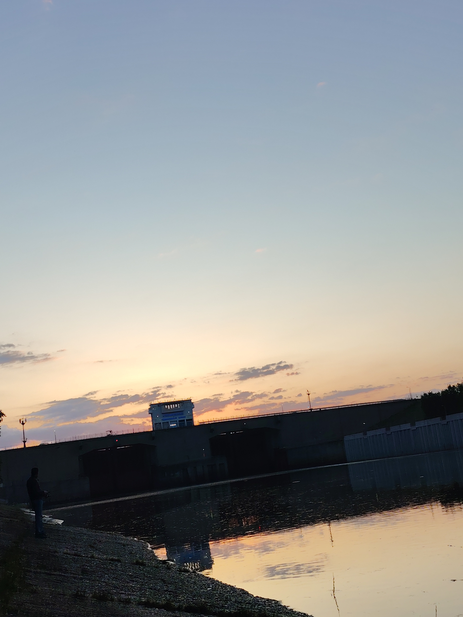 A wide-angle shot depicts a man fishing along a pebbled shoreline in Chaikovsky, Russia, at sunset. The water, reflecting the orange-tinged sky, meets the bank. A large, dark-colored bridge with a structure on top spans the water in the background. The sky transitions from pale blue to orange, with scattered clouds. The scene is peaceful, with the focus on the natural light and the silhouette of the man fishing.