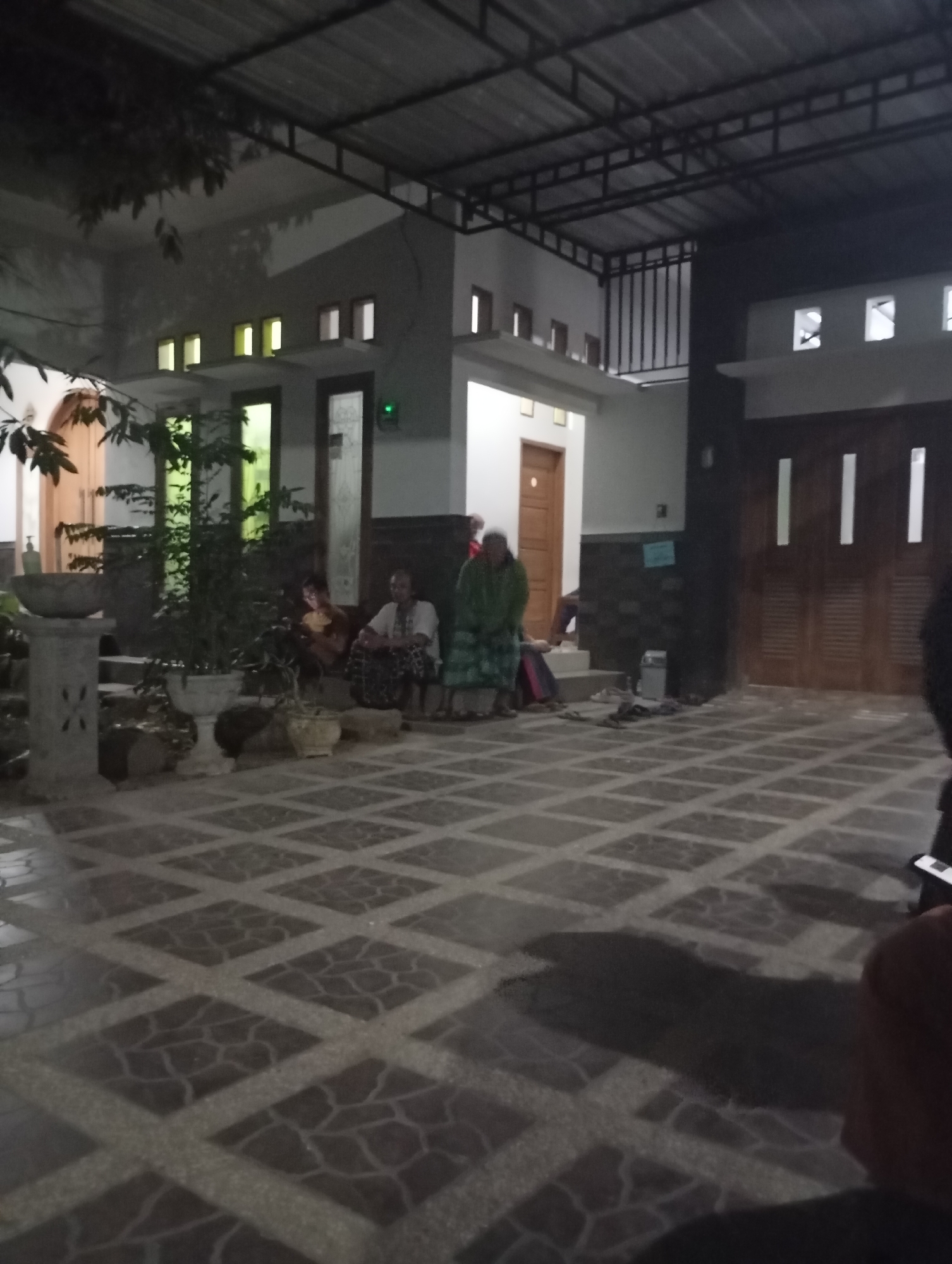 The image depicts an outdoor scene at night, likely in Banjarsari, Indonesia. The foreground features a patterned tiled floor. In the middle ground, several individuals are seated on the ground and on steps in front of a building. One person, dressed in a green garment, is standing. The building has a dark roof structure supported by metal beams. There are windows with internal lighting, and large wooden doors are visible. Plants in pots are placed around the area. The lighting suggests it is evening or night.