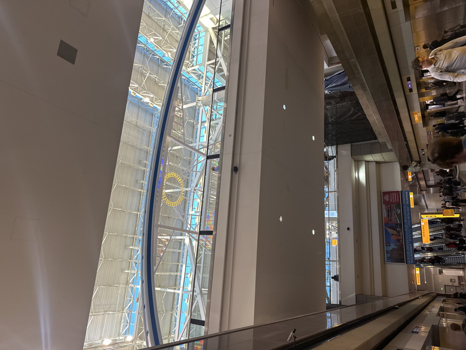 The image captures the interior of an airport terminal, showcasing a curved glass wall with structural supports and a circular yellow element. Adjacent to the glass structure is a broad, light-colored wall with small, evenly spaced lights. The scene extends into a bustling area with escalators, passengers, and signage. An overhead structure with linear elements creates a sense of depth and perspective.