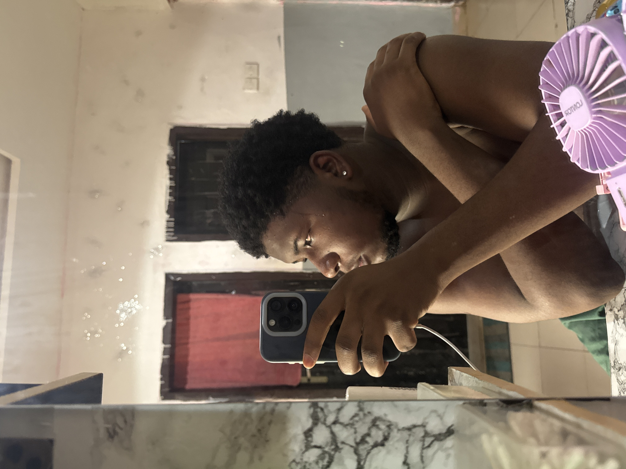 A person is taking a selfie in a bathroom. The person is facing down at a slight angle, holding a phone in their left hand. The phone is dark blue with a triple-lens camera. The person has dark hair and a beard, and is wearing an earring in their left ear. The person's right arm is folded across their chest. A purple fan is positioned on the right side of the frame. The bathroom has a marble countertop and a mirror. The wall behind the person is white with some smudges and water spots. There is a brown window frame visible behind the person. A red curtain is behind the window frame. The photo appears to be taken indoors. The city of Asipa, Nigeria, is not mentioned in the image.