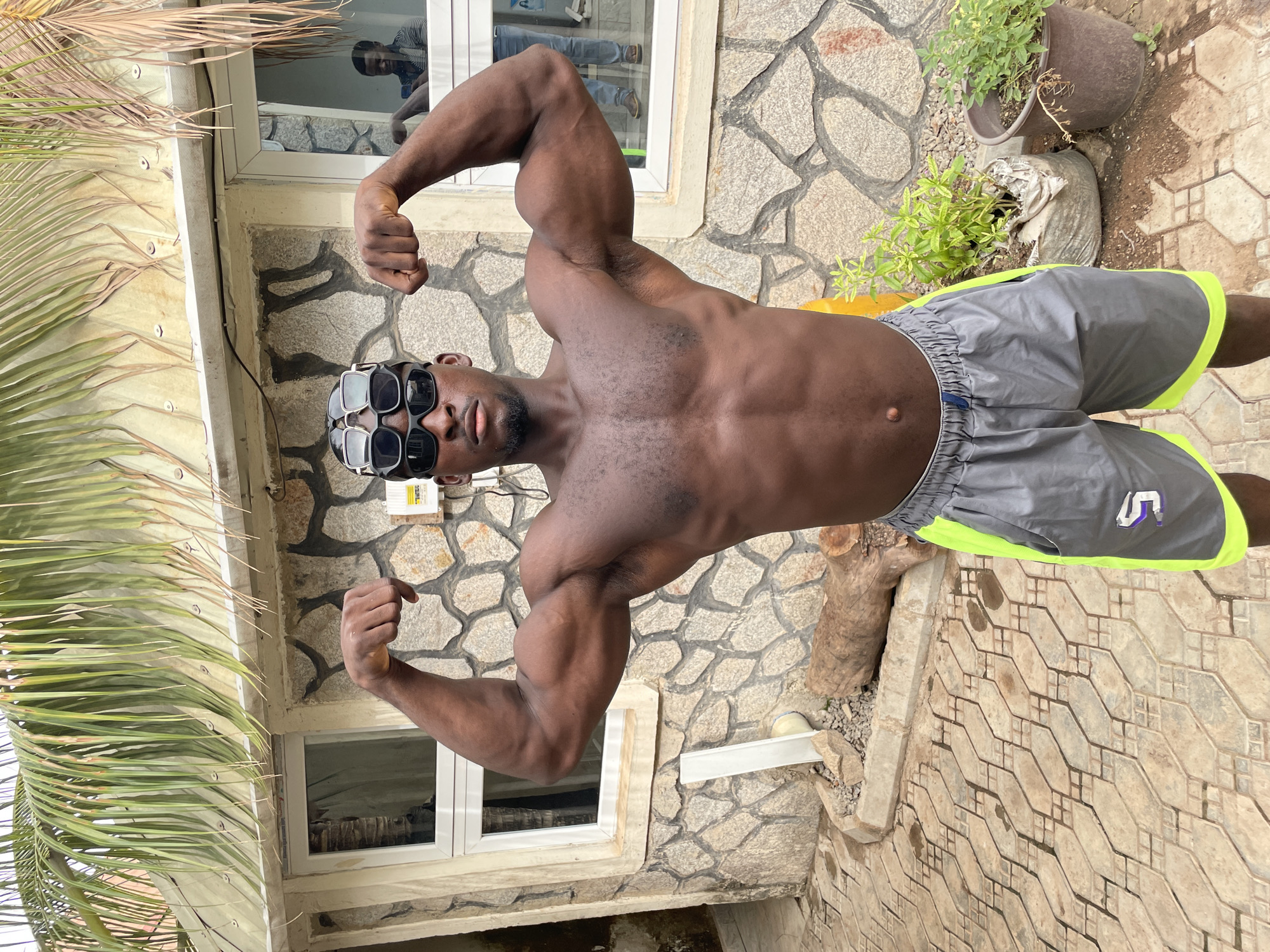 A dark-skinned, muscular man poses shirtless outdoors in Akwanga, Nigeria. He wears multiple pairs of sunglasses stacked on his face and grey shorts with a neon green trim. He is flexing his biceps. He is standing on a patterned stone patio with a building to his left with stone cladding and a window with a person reflected in it. A palm tree is visible to the left of the man, and potted plants are to the right. A drain pipe runs along the building wall near his shorts. A tree stump lies near the shorts. The image is rotated.