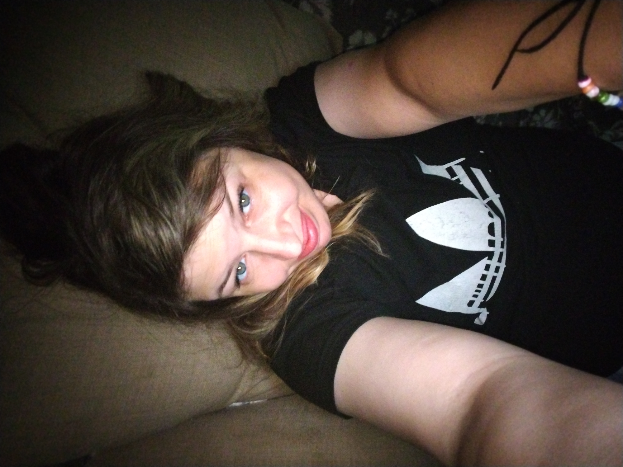 The image is a close-up, slightly blurry selfie of a young Caucasian woman lying down. She has brown hair and blue eyes. She is wearing a black t-shirt with a white logo on it. There is a beaded bracelet on her right wrist. The background appears to be a couch or soft surface, suggesting an indoor setting. The lighting is dim, creating shadows. There is no identifiable location in the city of Yaroslavl, Russia.