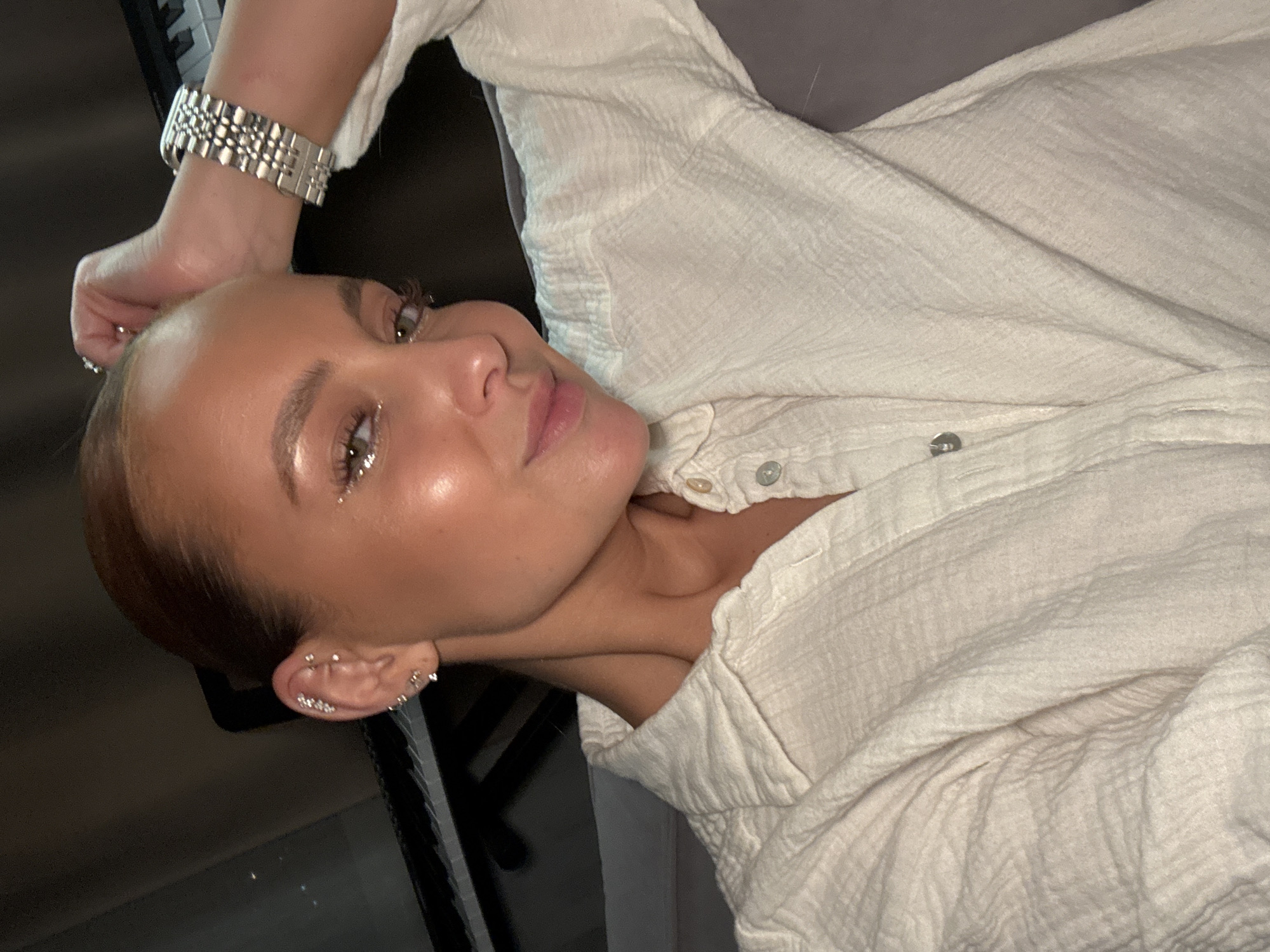 A fair-skinned woman with short, reddish-brown hair is lying on a neutral-colored surface. Her head is tilted towards the camera. She is wearing a light-colored, textured button-down shirt. She has glittery eye makeup. She wears a silver-colored watch on her left wrist and multiple earrings in her left ear. The scene includes a dark, partially visible object in the lower left, potentially part of furniture. The location is unknown and there is no information to suggest it is in Chaikovsky, Russia.